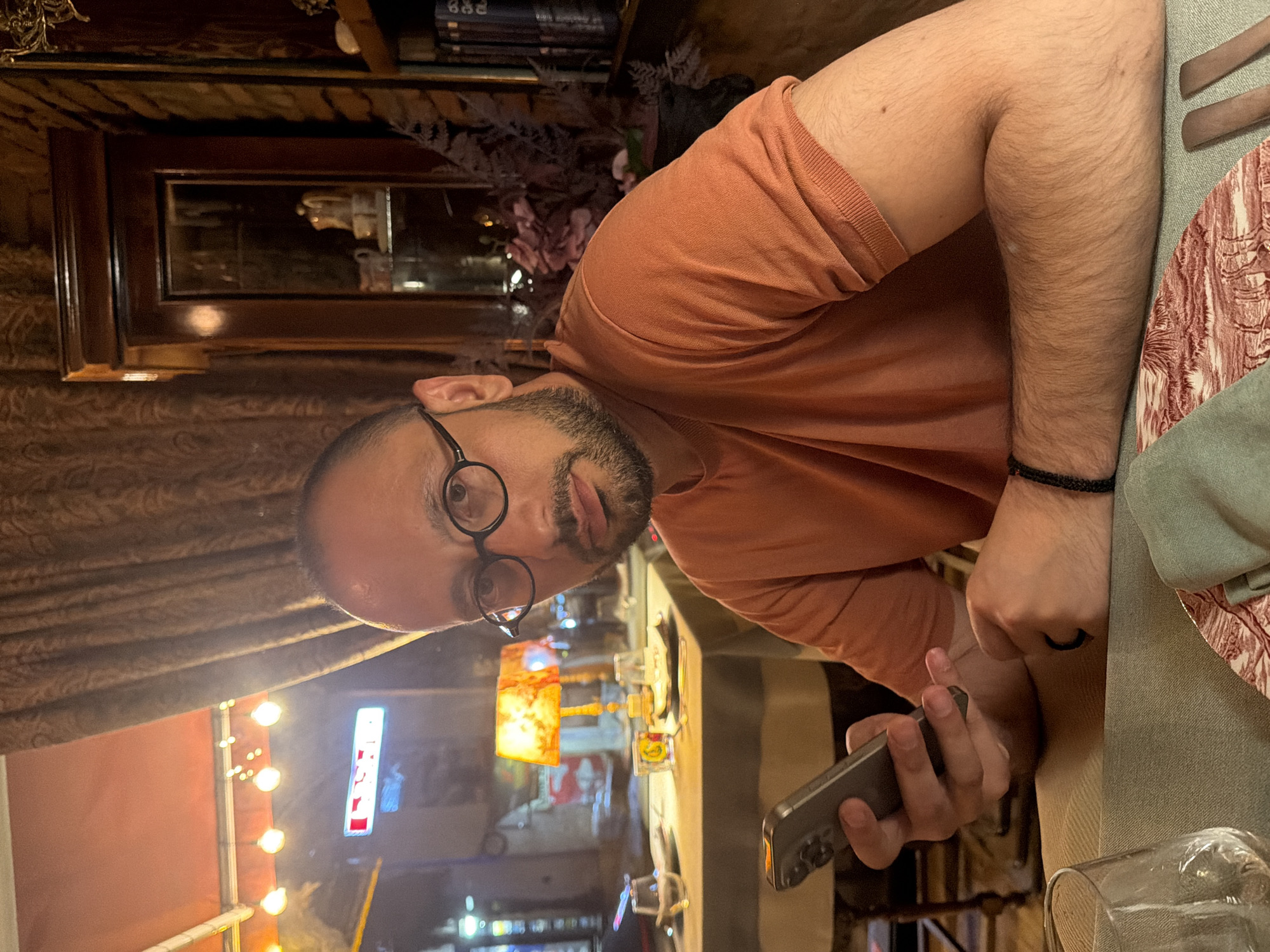A man with a bald head, glasses, and a beard is holding a cell phone at a table in what appears to be a restaurant or bar, likely located in the city of Tbilisi, Georgia. The outside is visible in the reflection of the glass table, showing lights, a red sign with white letters, and what seems to be a street scene at night. The man is wearing a peach-colored shirt and a black bracelet. The table is set with a green tablecloth, plates, utensils, and glassware. In the background, there is a shelf with books and decorative items.