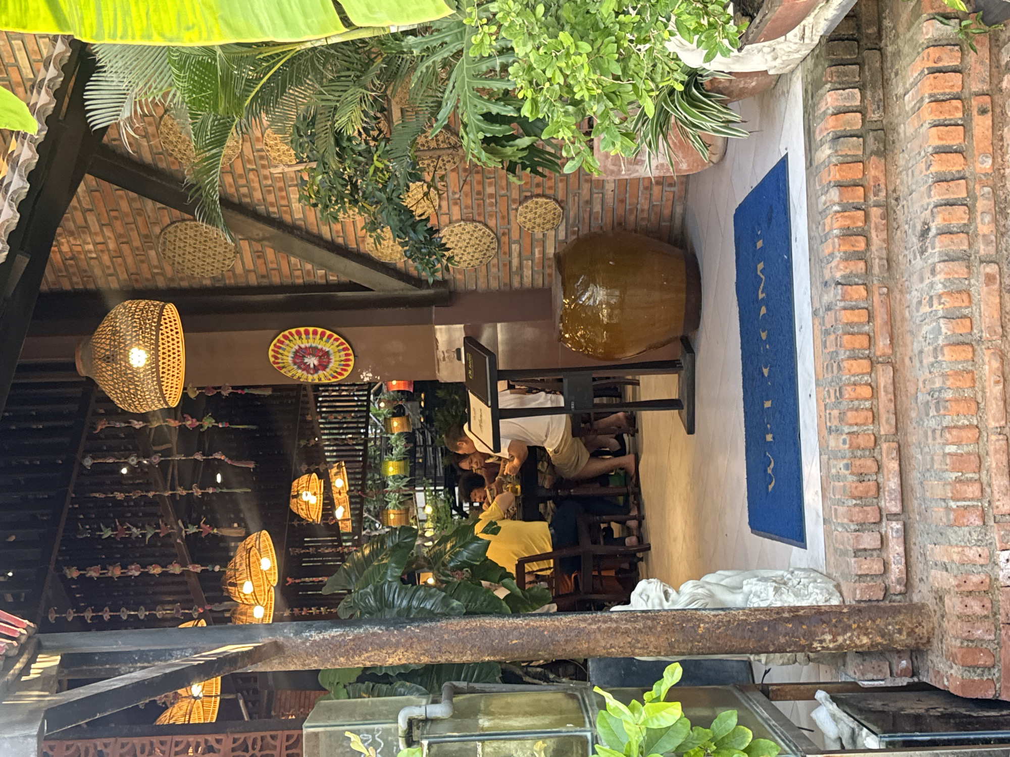 The image depicts the entrance to a restaurant in Nha Trang Ward, Vietnam. The entrance features a rustic design with exposed brick walls, wooden beams, and woven pendant lights hanging from the ceiling. Several potted plants, including large banana-like leaves and ferns, are strategically placed to enhance the ambiance. A large, brown ceramic pot sits beside the entrance, and a blue welcome mat with white lettering is visible on the floor.
Inside the restaurant, a group of people are seated at a table, appearing to be dining. One individual is wearing a bright yellow shirt, while others are in casual attire. The lighting is warm and inviting, casting a soft glow on the scene. The overall impression is that of a cozy and welcoming establishment.