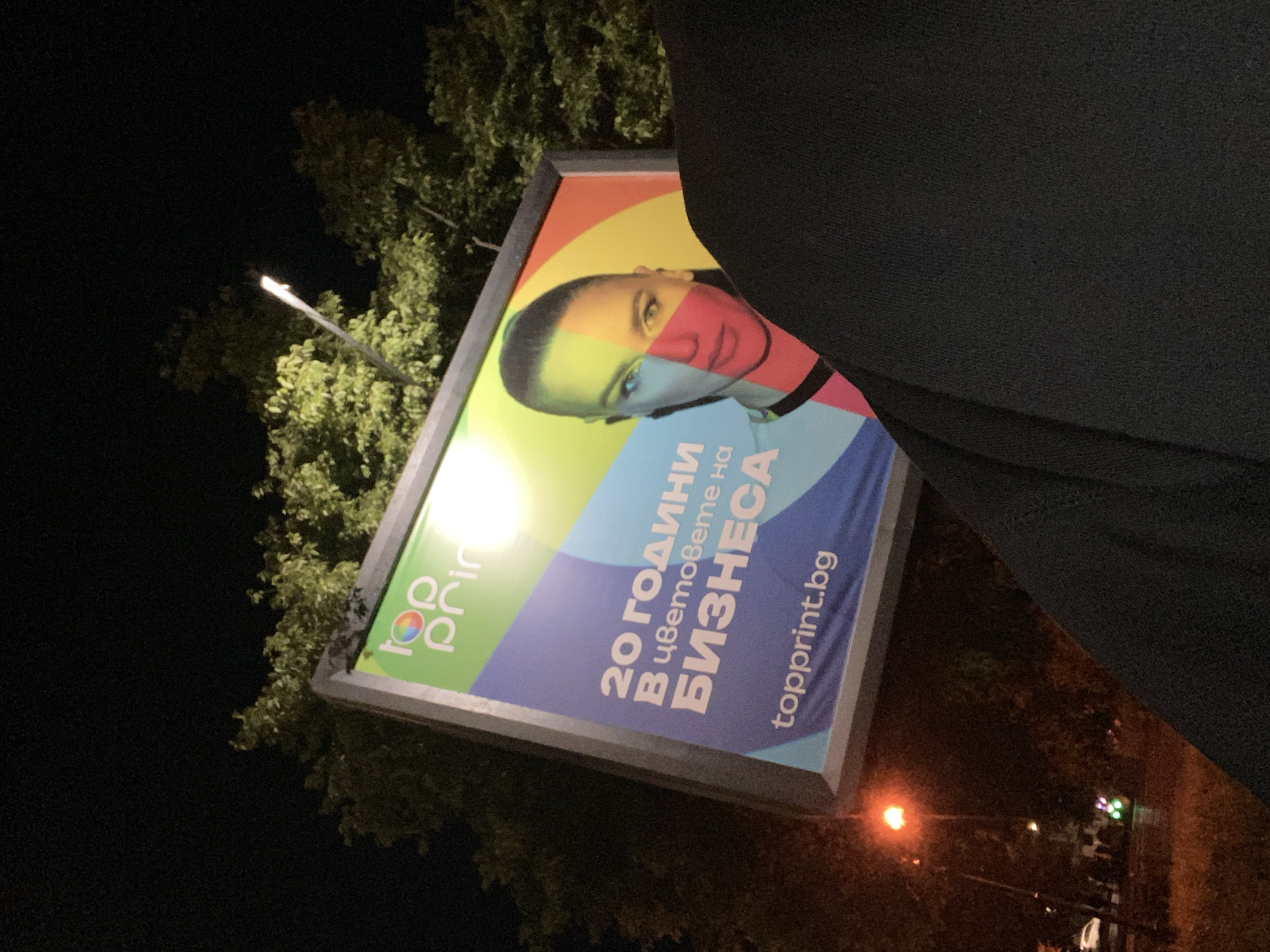 The image, taken at night, depicts a billboard advertisement for “top print” in Varna, Bulgaria. The advertisement features an image of a person whose face is divided into color segments, and the text “20 години в цветовете на БИЗНЕСА” along with the website “topprint.bg”. A streetlamp is visible in the upper left corner behind tree foliage. The foreground contains part of an individual wearing black clothing. In the background, on the right, there are additional streetlights and possibly other buildings. The billboard is situated among trees.