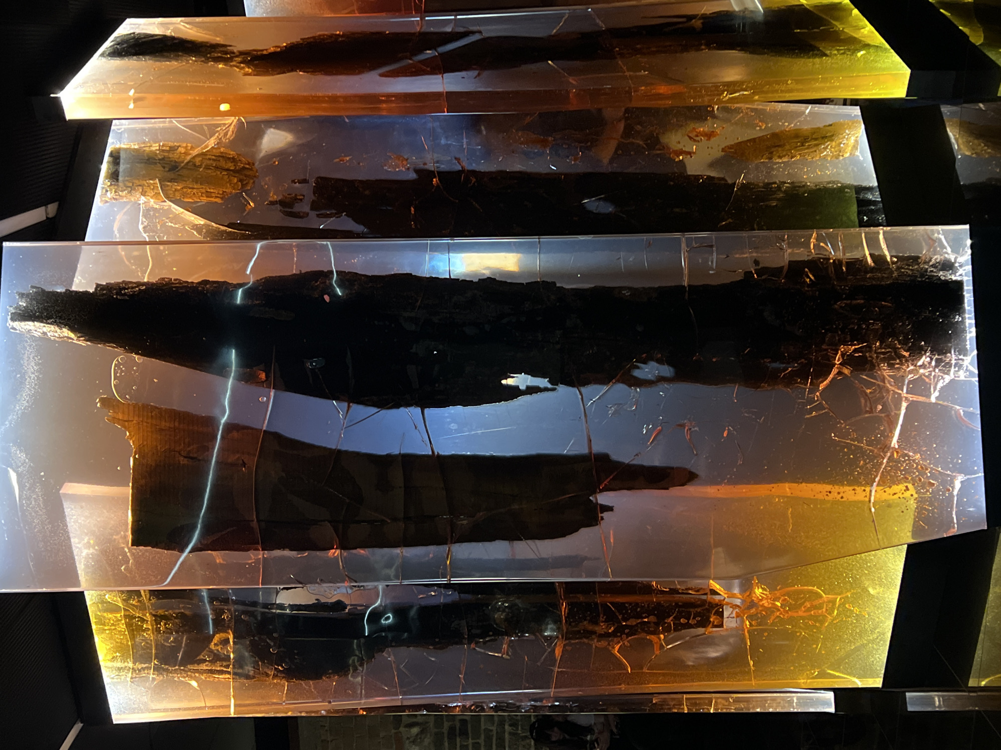 This image depicts an art installation in the city of Gdańsk, Poland. The installation features several tall, rectangular glass-like pillars, each encasing dark, rough organic material, possibly petrified wood or ancient artifacts. The pillars are illuminated from within and from below, with a warm, amber glow at the base and cooler tones higher up, creating a dramatic, almost ethereal effect. The surfaces of the pillars appear fractured and textured, with visible cracks and internal imperfections. In the background, a woman with dark hair is partially visible, seated and looking downwards, her presence adding a sense of scale and human interaction with the art. The environment appears to be an indoor exhibition space, with dark ceilings and textured brick walls visible in the far left background. The overall impression is one of historical preservation and artistic interpretation, with the materials seemingly suspended in time within the transparent medium.