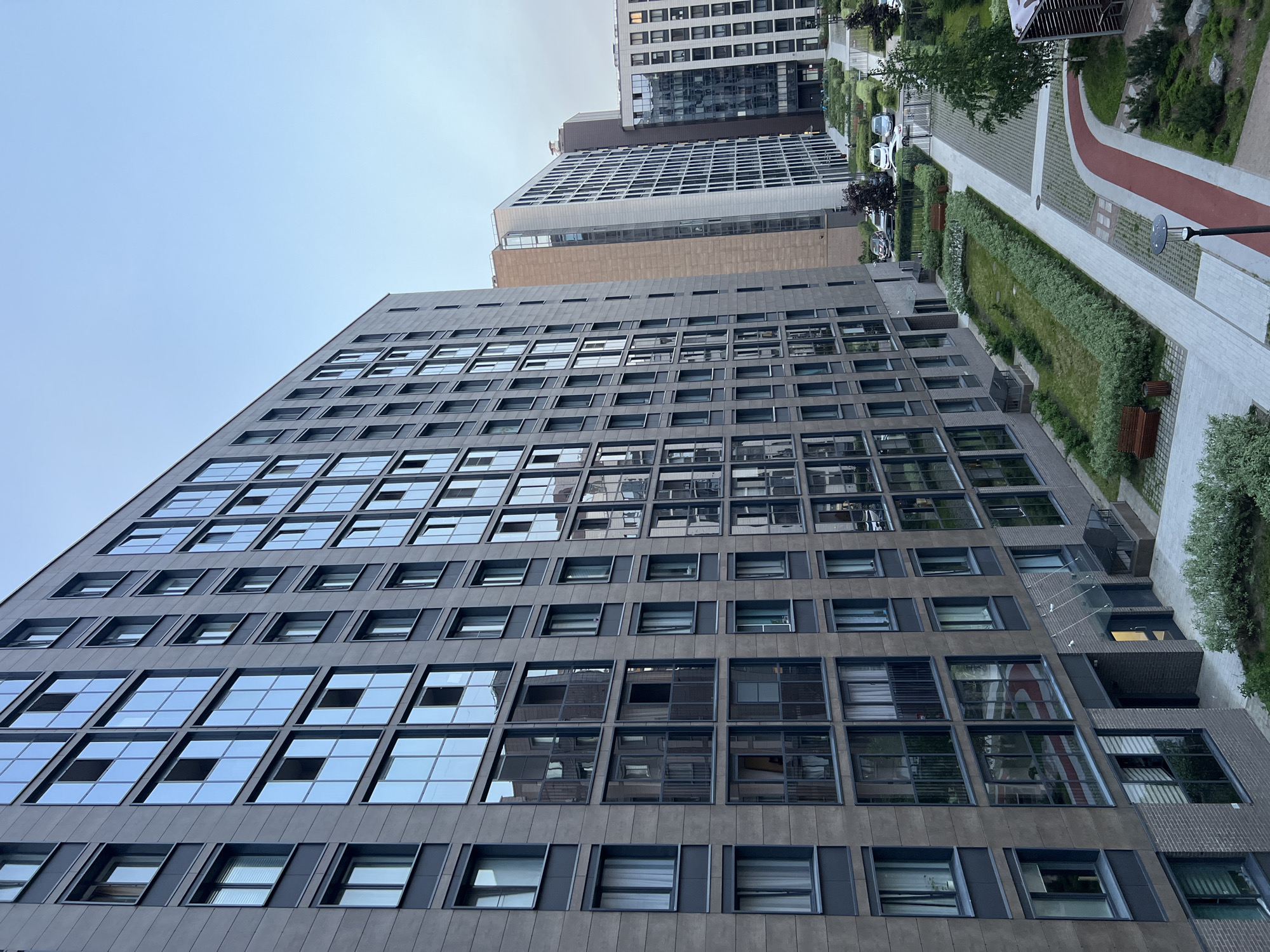 The image depicts a modern residential complex in the Chistoe Nebo residential complex, Primorsky District, Saint Petersburg, Russia. The scene is observed from an elevated perspective, likely from an upper floor of one of the buildings.
In the immediate foreground and left, a multi-story residential building dominates the frame, presenting a facade composed of dark grey/brown panels. This building features numerous rectangular windows, many of which are full-height and reflect the light blue sky, surrounding buildings, and parts of the courtyard below. Some windows have internal curtains or blinds visible. The lower levels of this building incorporate a darker brick cladding near multiple ground-level entrances, each protected by a small, translucent canopy.
To the right of the foreground building, a series of other modern high-rise residential structures are visible in the middle ground and background. One prominent building has a lighter grey/white facade with a grid pattern of windows, interrupted by a vertical section of beige/tan cladding. Further right, another building exhibits a darker, more reflective glass facade.
The ground level between the buildings features a meticulously landscaped courtyard. Light grey paved walkways traverse the area, bordered by neatly maintained green lawns and various bushes forming hedges. Several wooden benches are placed along these paths. A distinctive red-surfaced path, designed for walking or jogging, curves through the courtyard. Overhead street lamps are integrated into the design. In the distance, beyond a low fence and gate, a paved parking area contains several parked automobiles, including a white and a dark-colored sedan. No individuals are visible within the frame, and the scene appears static under a clear, light blue sky with minimal cloud cover.