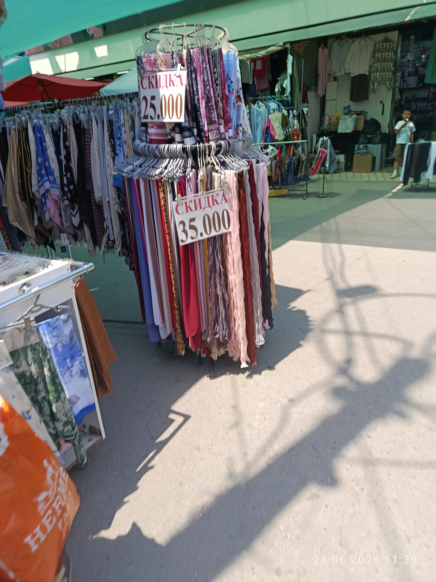An outdoor market stall displays an assortment of scarves for sale. The scarves are hung on a circular rack, with some also on hangers. Two signs are visible, both reading "СКИДКА" (discount) followed by a price in Russian currency. One sign shows "25.000" and the other "35.000". The stall is set up under a green awning, and the ground is paved. In the background, other stalls and a young boy wearing a white t-shirt and shorts are visible. The time stamp "28.06.2026 11:39" is overlaid on the bottom right of the image.