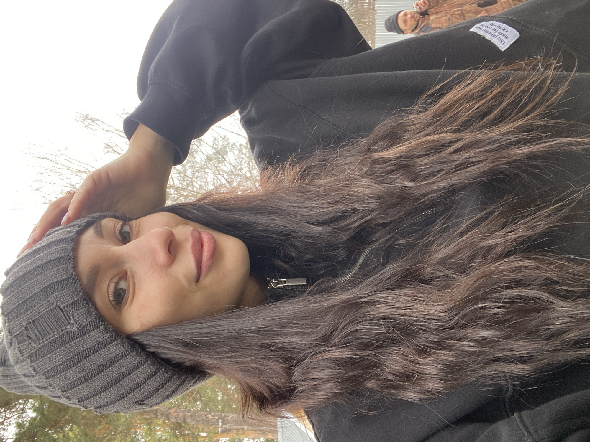 A person wearing a gray beanie and a black hoodie is looking at the camera. The hoodie is zipped up. The person's hair is dark brown and wavy. The background is a blurred out landscape of trees and a pale sky. Another person is visible in the upper right, wearing a hat, with clothing featuring a camouflage pattern. A white label is attached to the hoodie near the second person. The location may be Chaikovsky, Russia, based on the prompt, but there is no verifiable visual evidence in the image to confirm this.