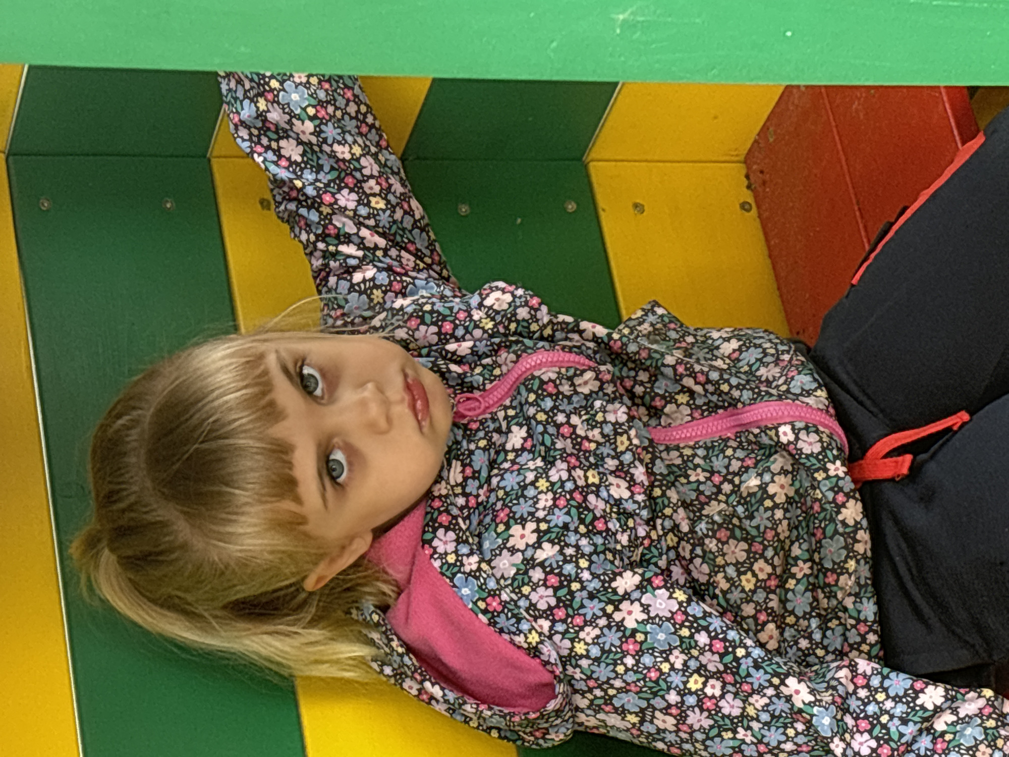 A young Caucasian girl with blonde hair in a ponytail and bangs is sitting in a brightly colored structure. The structure has alternating green and yellow horizontal panels on the back wall and a red bench seat. The girl is wearing a floral-patterned hooded jacket with a pink lining and a pink zipper, and black sweatpants with a red drawstring. Her expression is glum. The background is a corner of the structure, with green and yellow panels. This scene appears to be an indoor play area or a similar recreational space. The location is identified as Novye Chernitsy, Russia.