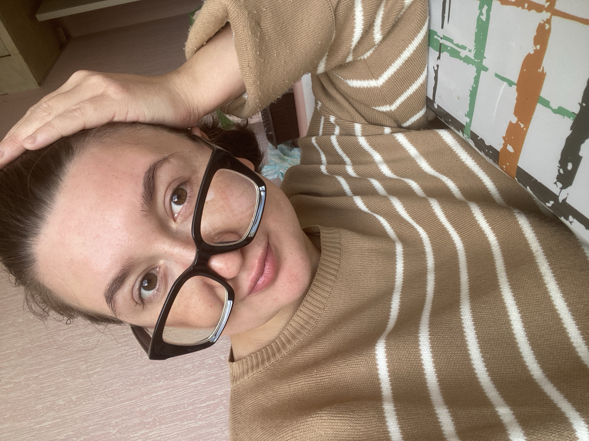 A woman is pictured from the shoulders up in a close-up, casual portrait. She has brown hair pulled back, is wearing black-rimmed glasses, and a tan and white striped sweater. Her right hand is resting on the top of her head. She is looking towards the camera with a neutral expression. The background is a pale pink textured wall. The lower right portion of the image shows a tabletop with a checkered pattern of orange, green, and brown lines on a white background. No specific location within Chaikovsky, Russia is identifiable.