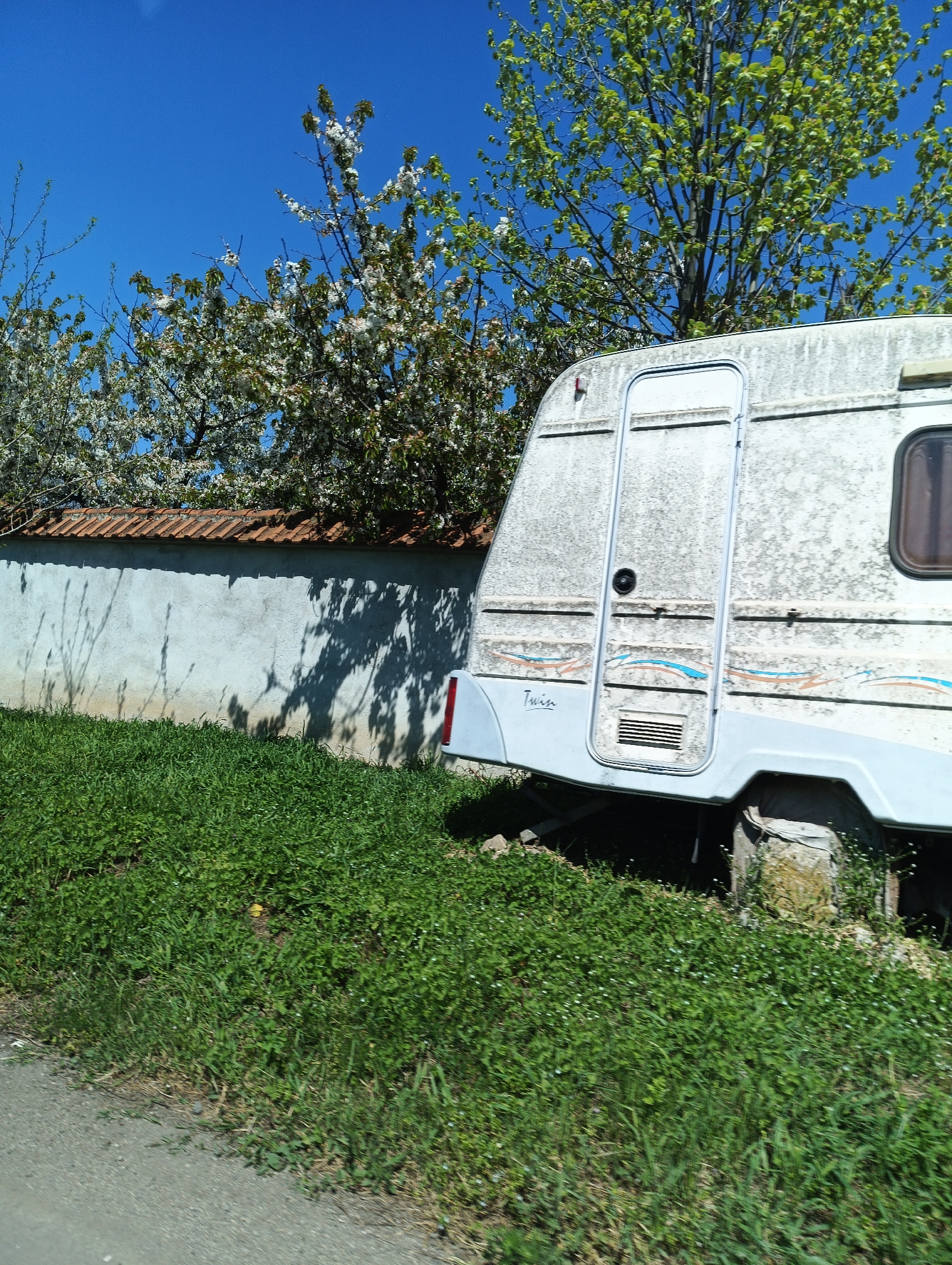The image shows a dilapidated white caravan with the word "Twin" on its side, parked in Straldzha, Bulgaria. The caravan is adjacent to a low, white wall with a red-tiled top. Trees with white blossoms are visible behind the wall. The caravan is partially obscured by overgrown green grass. A paved road is visible in the lower left corner. The sky is clear and blue.
