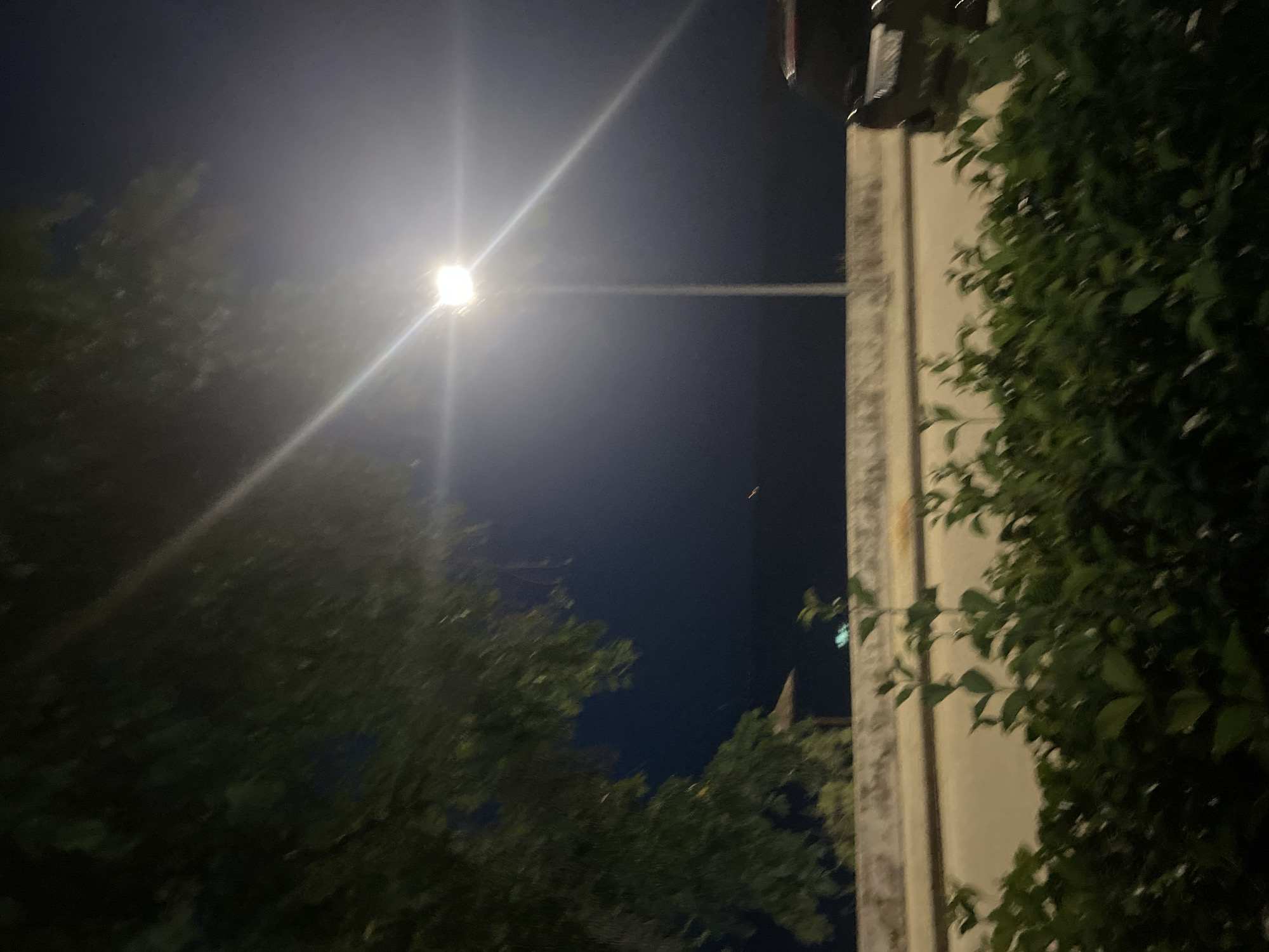 This media file captures a night scene in Nea Karvali, Greece. The primary subjects are a street lamp illuminating the dark sky and surrounding trees. The bright light of the lamp creates strong lens flares that dominate the upper left quadrant of the image, with visible light rays extending outward. To the right, a building's facade, likely made of concrete or stone, is partially visible, with dense green foliage climbing up its side. The foliage is lush and appears healthy.
The scene depicts a quiet night, possibly in a residential area or park, given the presence of trees and a building. The deep blue of the night sky suggests it is well after sunset. There is no indication of specific weather conditions, though the bright light from the lamp suggests clear skies rather than fog or heavy cloud cover.
Notable details include the contrast between the bright artificial light and the dark natural elements of the trees and sky. The way the light interacts with the foliage creates areas of deep shadow and illuminated leaves. In the distance, a faint point of light is visible in the sky, which could be another light source or an aircraft. A small, pointed structure, possibly part of a building or a decorative element, is faintly visible behind the trees on the right side of the frame.
No people are visible in the image, and no specific activity or event is clearly taking place, creating a sense of stillness and quiet. The overall mood is one of peaceful observation of an urban nightscape illuminated by a single strong light source. There is no visible text within the image.