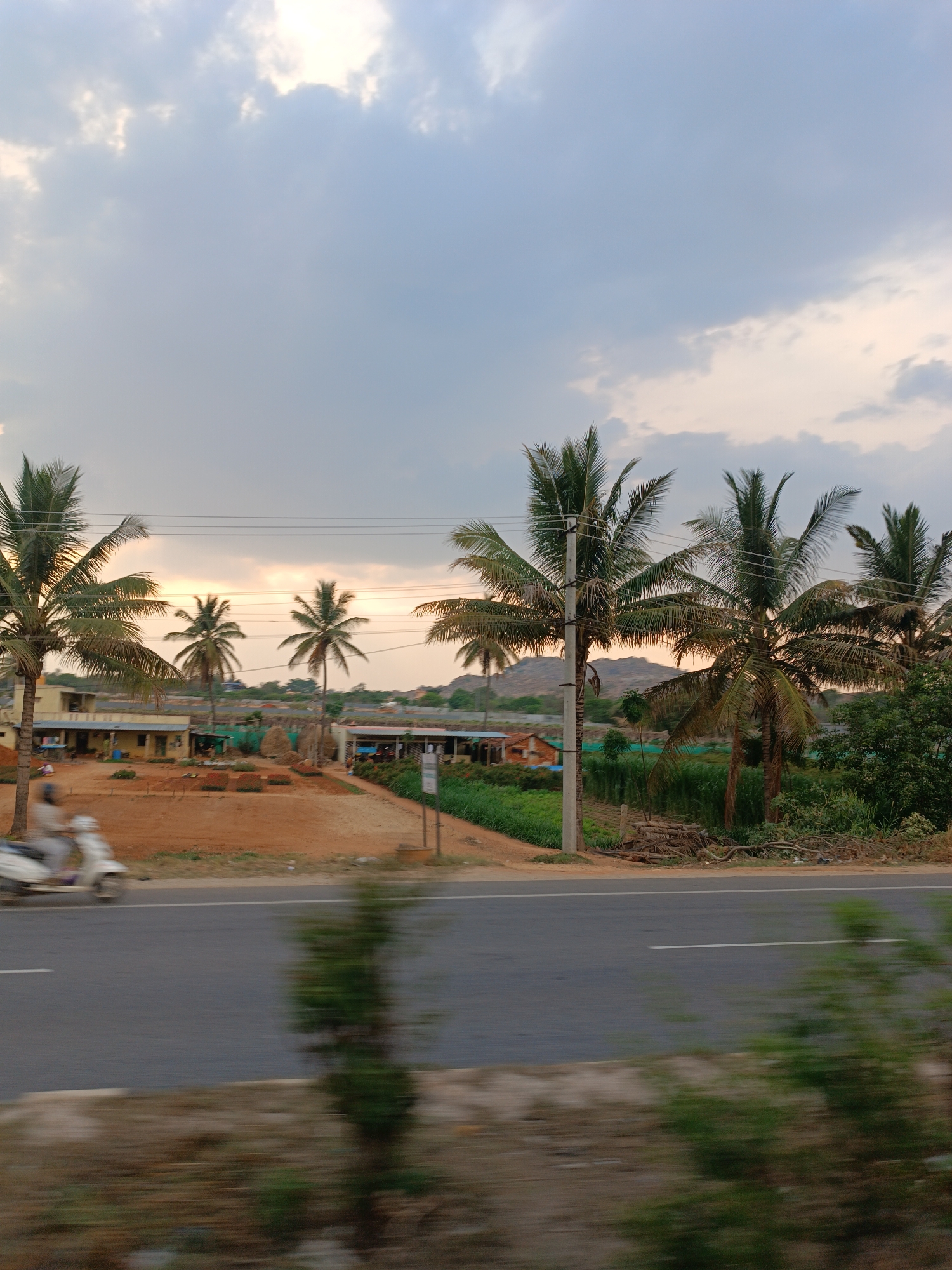The photograph shows a landscape in Attibele, India, under a partly cloudy sky. A road runs horizontally across the foreground, blurred due to motion, with low vegetation along its edge. Behind the road, the ground is reddish, possibly due to the type of soil. Several palm trees are spaced across the scene, with utility poles among them. Beyond the trees are buildings, some of which appear to be houses or shops. Behind the buildings are low, rocky hills. A scooter is moving on the road, adding a dynamic element to the otherwise still scene. The lighting suggests either dawn or dusk.