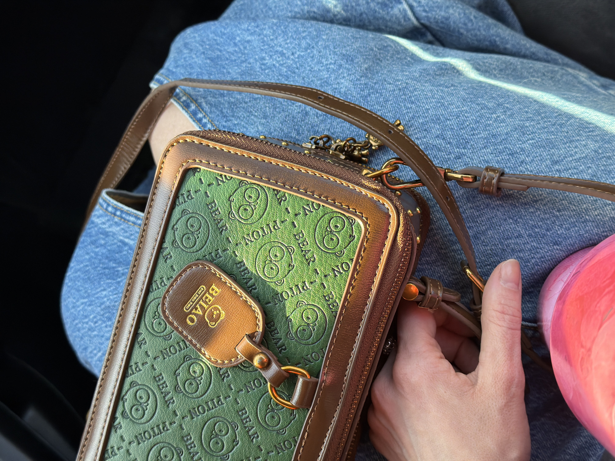 A medium shot captures a brown and green purse or small handbag with a "BEIAO" tag, set against a background of blue denim fabric. The purse is held by a hand with visible fingernails and wrinkles, positioned near what appears to be a pink water bottle or similar item. The bag features a repeating pattern of stylized bear images and the words "PITON" and "BEAR." The environment suggests an indoor setting, potentially within a car or similar confined space, given the intimate proximity of the objects and the visible person. No indication of Perm, Russia is found in the image.