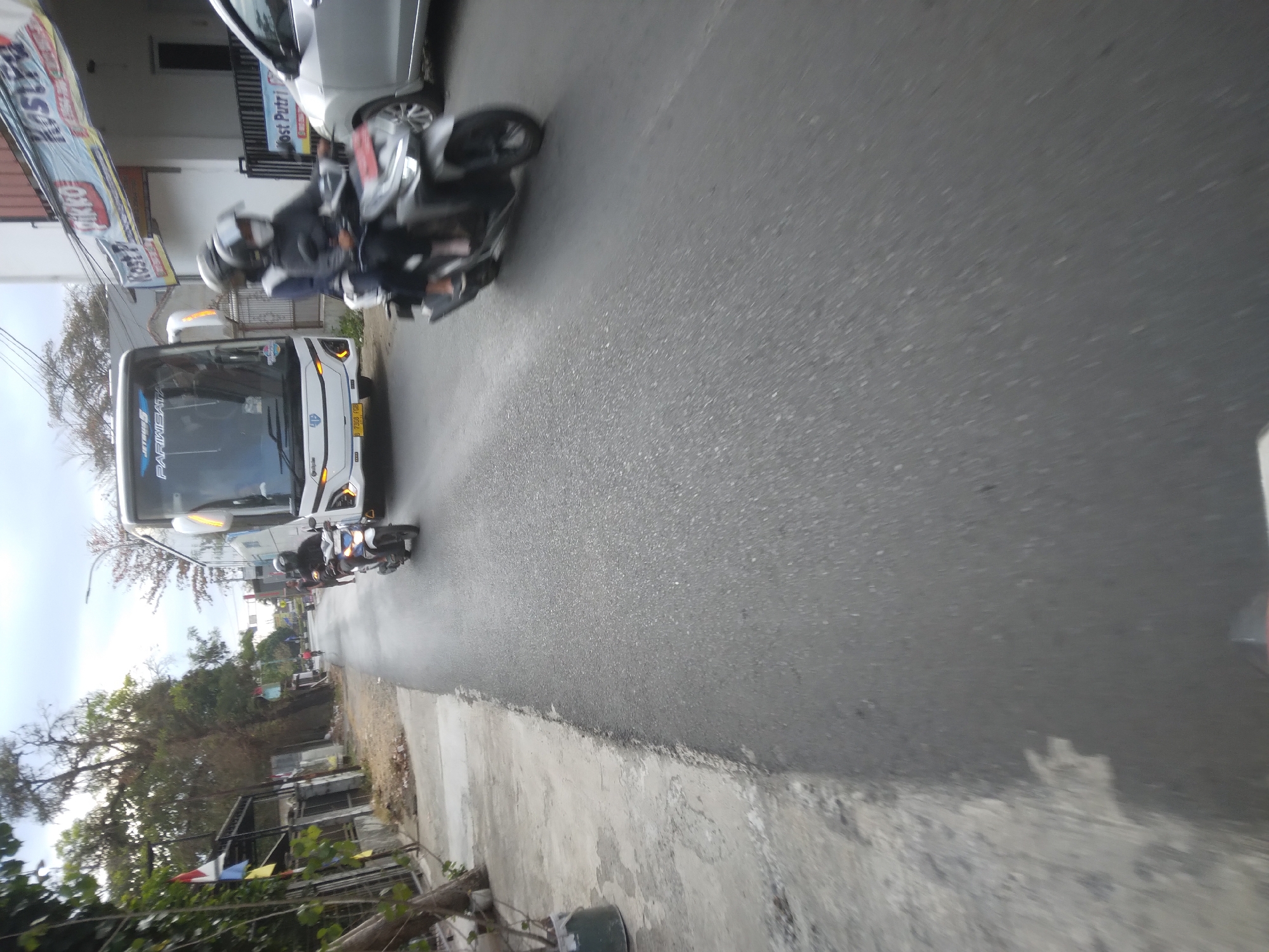The image depicts a street scene in Desa Pamijen, Kecamatan Sokaraja, Indonesia. A white tour bus with "JETBUS" and "PARIWISATA" visible on its front is driving on the right side of the road. To the right of the bus, a silver car is partially visible. In front of the bus and car, two motorcycles are traveling. The first motorcycle, closer to the bus, is ridden by a person wearing a black helmet and dark clothing. The second motorcycle, further ahead, carries two individuals, both wearing helmets. The motorcycle itself is silver with black accents. The road is paved and appears to be wet, with visible asphalt texture. On the left side of the road, there is a concrete sidewalk with some greenery and trees. Buildings with signs, including one that reads "KOST Putri," are visible along the roadside. The sky is overcast. The perspective suggests the photo was taken from a moving vehicle, likely another motorcycle, as indicated by the blurred foreground.