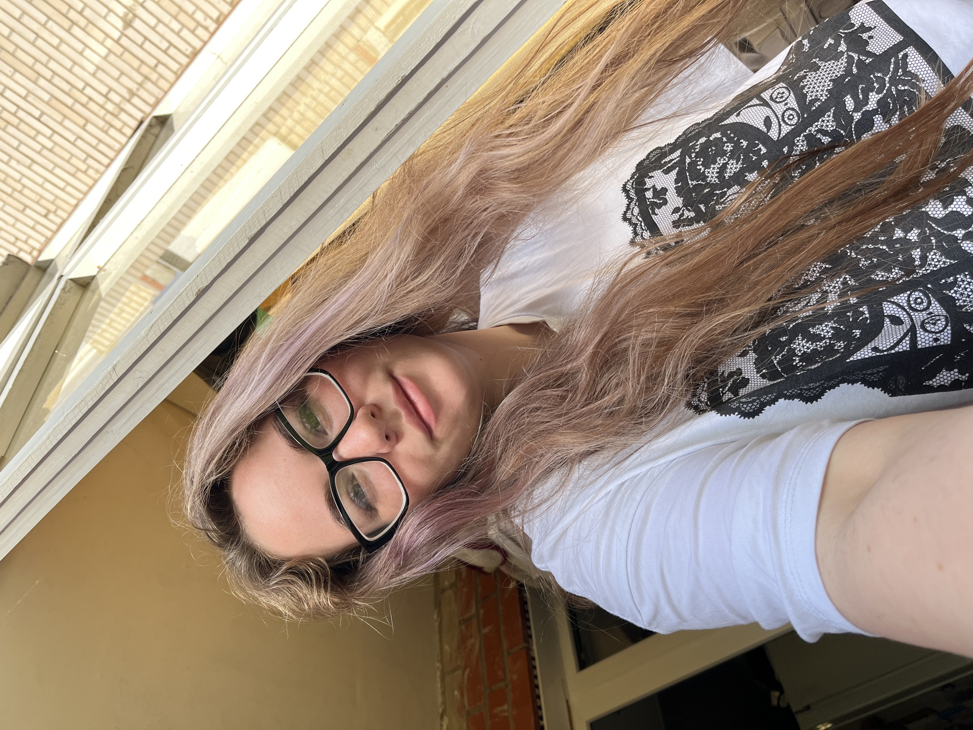 A woman with long, light brown and purple hair, wearing black-framed glasses and a white t-shirt with black lace detailing on the chest, is photographed from a high angle. The background includes a partially visible brick building, likely exterior siding. Architectural elements like window frames are also visible. The location may be Chaikovsky, Russia.