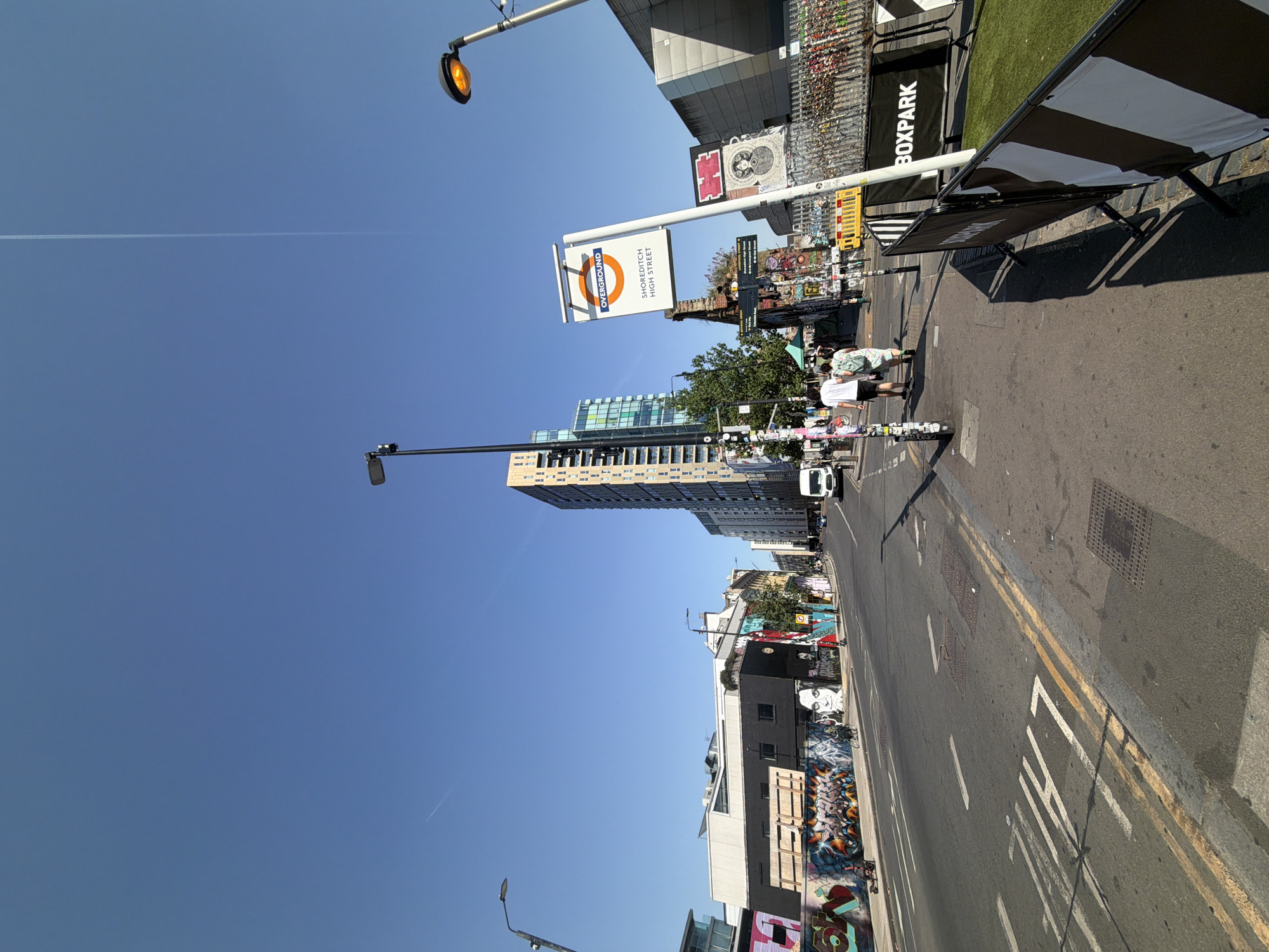 The image shows a street scene in Shoreditch, London, United Kingdom, with a view of Shoreditch High Street. The photograph is oriented vertically. In the upper right quadrant is a sign for "BOXPARK" along with a portion of a temporary barricade. Adjacent to the BOXPARK sign is the Shoreditch High Street Overground station signage. In the midground, two people are walking down the pavement. A tall, modern building is visible further down the street. Graffiti adorns many of the buildings along the street. The sky is clear and blue. Street lighting and signage poles are present. The street has double yellow lines and road markings. A white van is visible in the distance along the road.