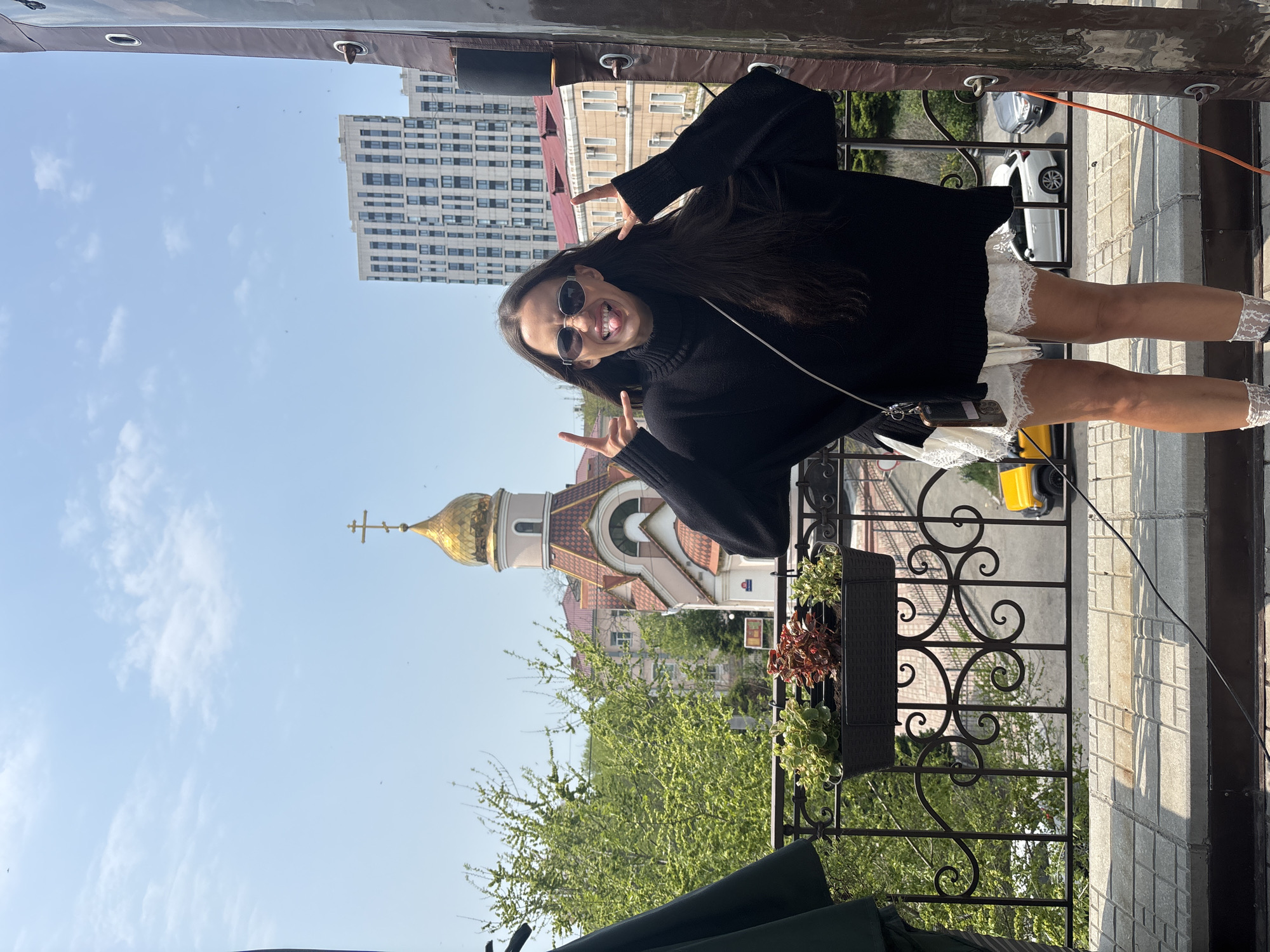 A woman stands on a balcony, making a peace sign with both hands. She has long dark hair, wears sunglasses, a black sweater, and a white dress. A purse hangs from her shoulder. Behind her is a cityscape, including a church with a gold dome topped with a cross and a tall, modern apartment building. Green trees and bushes fill the space below the balcony. The balcony has a black wrought-iron railing with flower boxes. The scene appears to be in Vladivostok, Russia, based on the architecture and location of the church and apartment building. The sky is blue with some white clouds. A yellow toy car is visible on the street in the background.