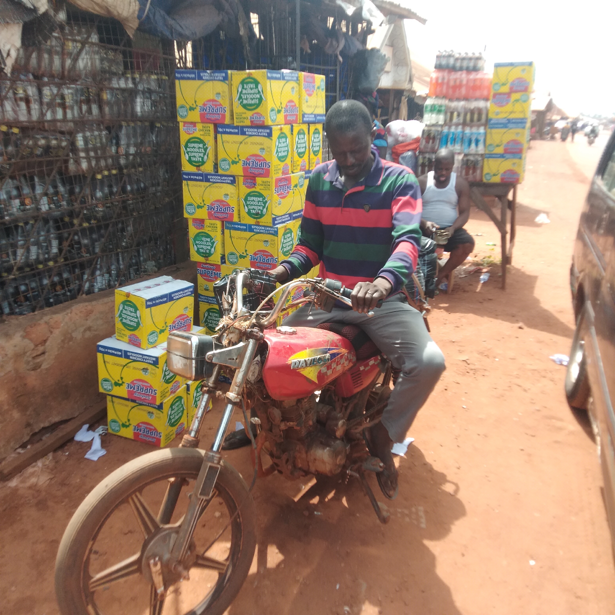 A man in a horizontally striped shirt and grey pants sits on a red Daylong motorcycle in front of a roadside stall in Ejule, Nigeria. The stall displays stacks of yellow boxes labeled "Supreme Noodles" and metal cages containing bottles. A second man sits in the background, and a brown vehicle is parked on the right edge of the frame. The ground is reddish-brown dirt.
