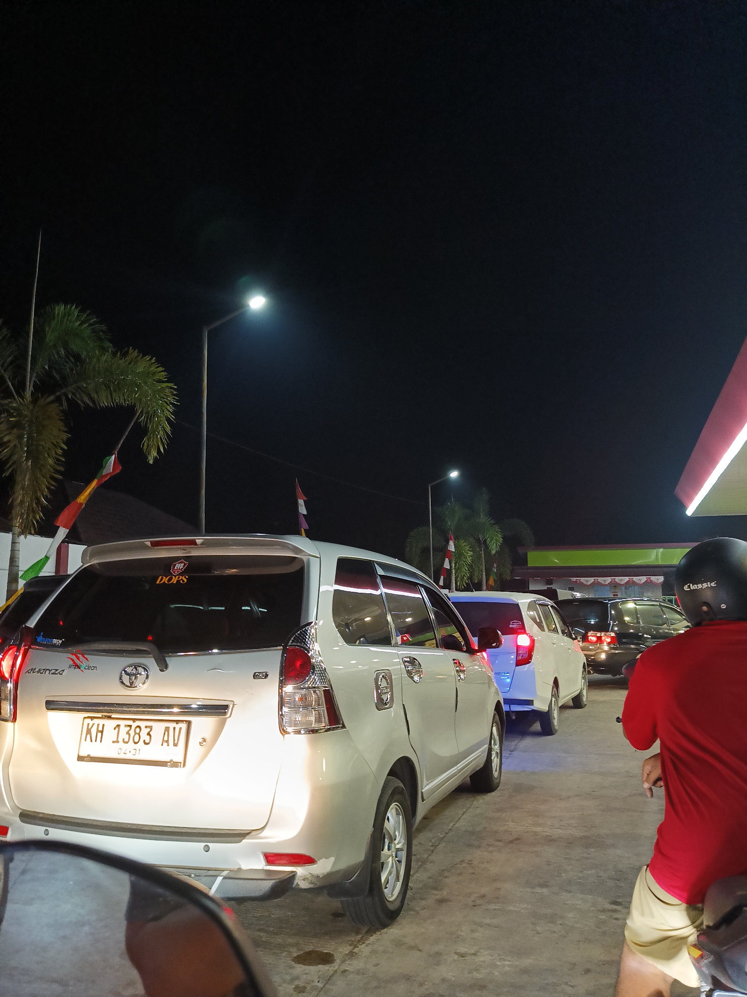 In Palangka Raya, Indonesia, at nighttime, a queue of vehicles is forming at a gas station, likely due to fuel shortages. The scene captures a silver Toyota Avanza with license plate KH 1383 AV in the foreground, its rear illuminated by bright lights. A person on a motorcycle, wearing a red shirt and beige shorts, is positioned to the right, facing away from the camera. Behind the Avanza, a white car is visible, followed by a black car. Palm trees and flags, possibly national flags, are present on the left side of the image, suggesting a public space. The overall atmosphere is one of congestion and waiting, indicative of a common scenario in many Indonesian cities.