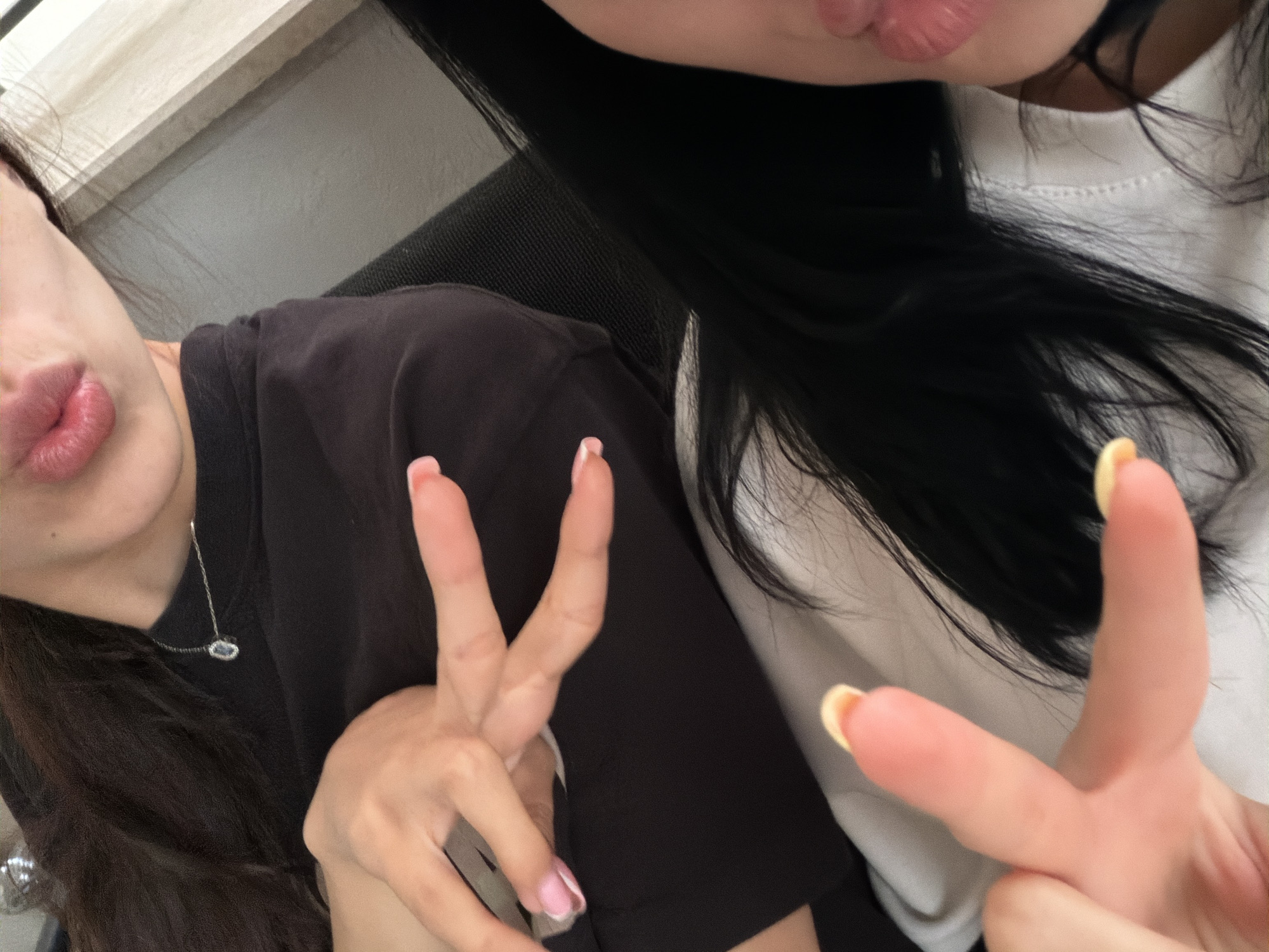 This is a close-up, over-the-shoulder selfie featuring two young women. The woman in the foreground is wearing a black t-shirt and a delicate silver necklace with a blue pendant. She has long, dark hair and her face is partially visible, showing her lips. She is holding up two fingers in a peace sign. The woman in the background is wearing a white t-shirt and also has long, dark hair. She is also holding up two fingers in a peace sign. Both women have manicured nails. The lighting suggests an indoor setting, possibly during the day. There is no visible text in the image. The overall mood appears casual and friendly.
