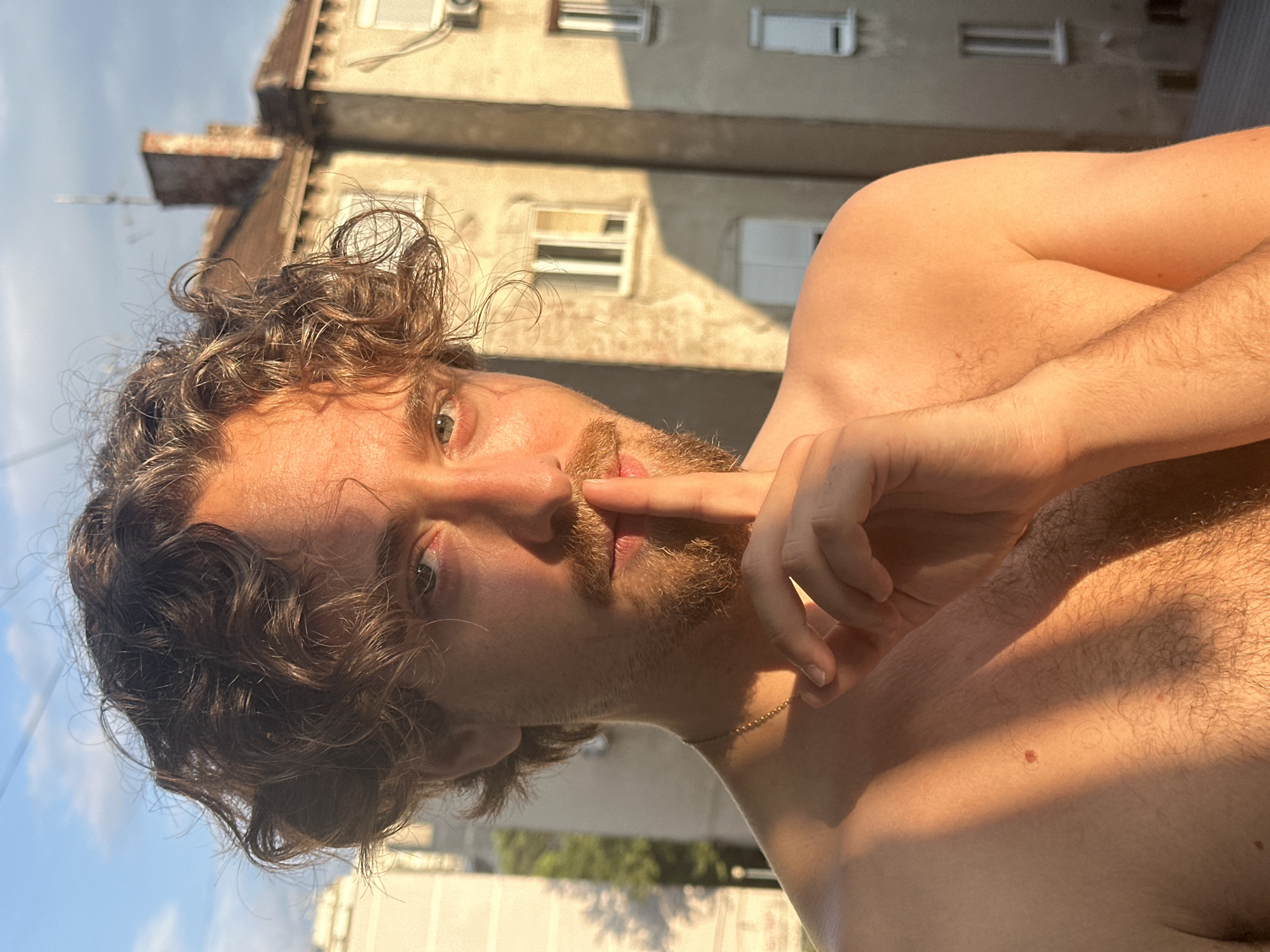 A close-up, low-angle, outdoor selfie of a young man with curly brown hair and a beard, set against a backdrop of apartment buildings and a blue sky. The man is shirtless, with his right hand raised to his mouth, and his gaze is directed towards the camera. The sunlight casts a warm glow on his skin, suggesting it is either early morning or late afternoon. The buildings in the background appear to be typical residential structures, common in urban environments. The overall impression is casual and intimate, possibly capturing a moment of reflection or playfulness. The image is oriented vertically. There is no visible text in the image.