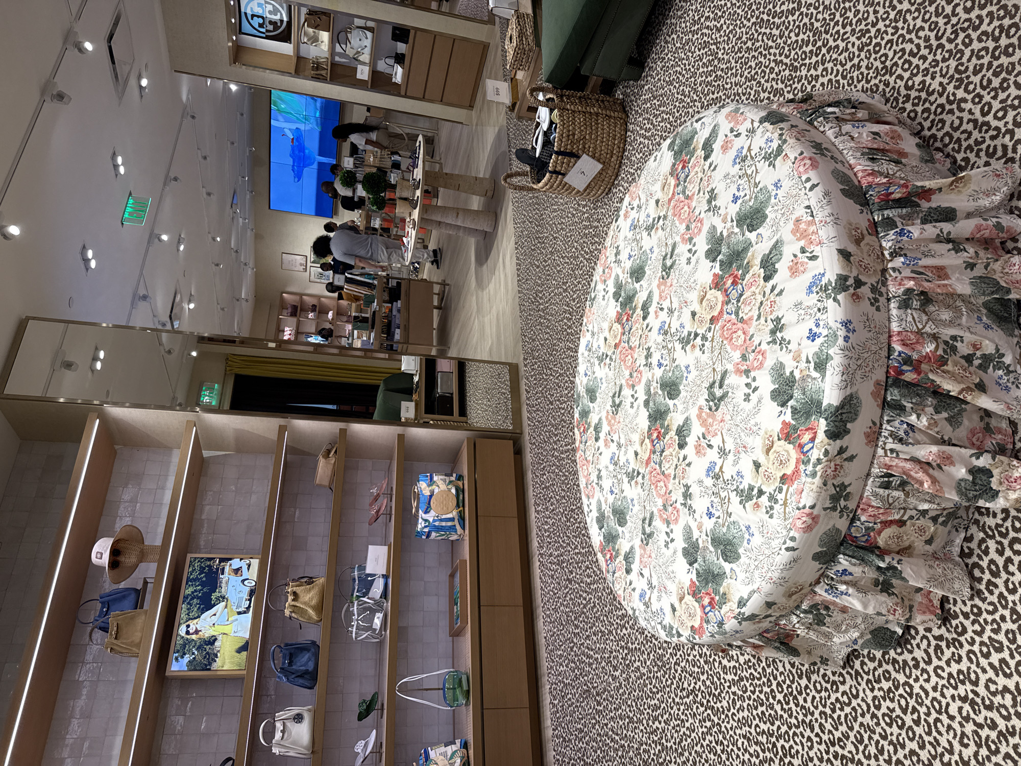 The image shows the interior of a store in Sunrise, United States of America. A round ottoman with a floral pattern and a ruffled edge is the prominent feature, sitting on a floor covered with an animal print rug. Near the ottoman, there's a wicker basket with shoes inside.
In the background, there are shelving units displaying various items like handbags, hats, and framed pictures. A large television screen hangs on the wall, and a group of people can be seen standing near it.