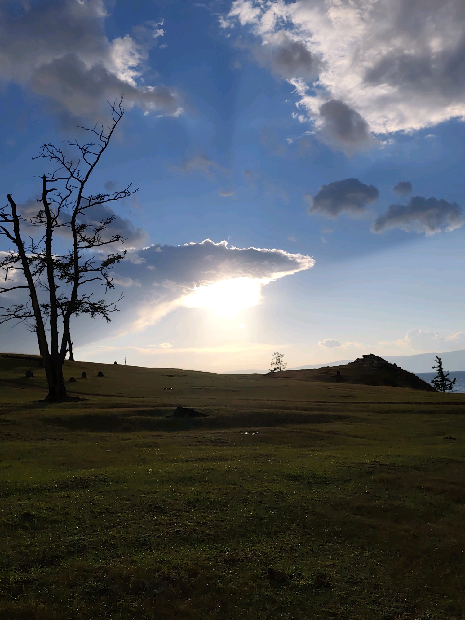 A vertical, low-angle, eye-level shot captures a landscape scene at dusk in Kharantsy, Russia. The foreground is a field of dark green grass, with some patches of yellow and brown. In the midground, a rolling hill is dotted with several small, dark mounds, possibly rocks or animal burrows. To the left, a tall, silhouetted dead tree stands prominently, its bare branches reaching towards the sky. In the distance, on the right side of the frame, a rocky outcropping rises from the land, topped with a lone tree. Beyond that, the calm water of a lake is visible, and further in the background, a hazy mountain range fades into the horizon. The sky is a dramatic mix of deep blue and white clouds, with the sun partially obscured behind a large, white cloud formation, casting rays of light downwards. The overall lighting suggests the transition from late afternoon to evening.