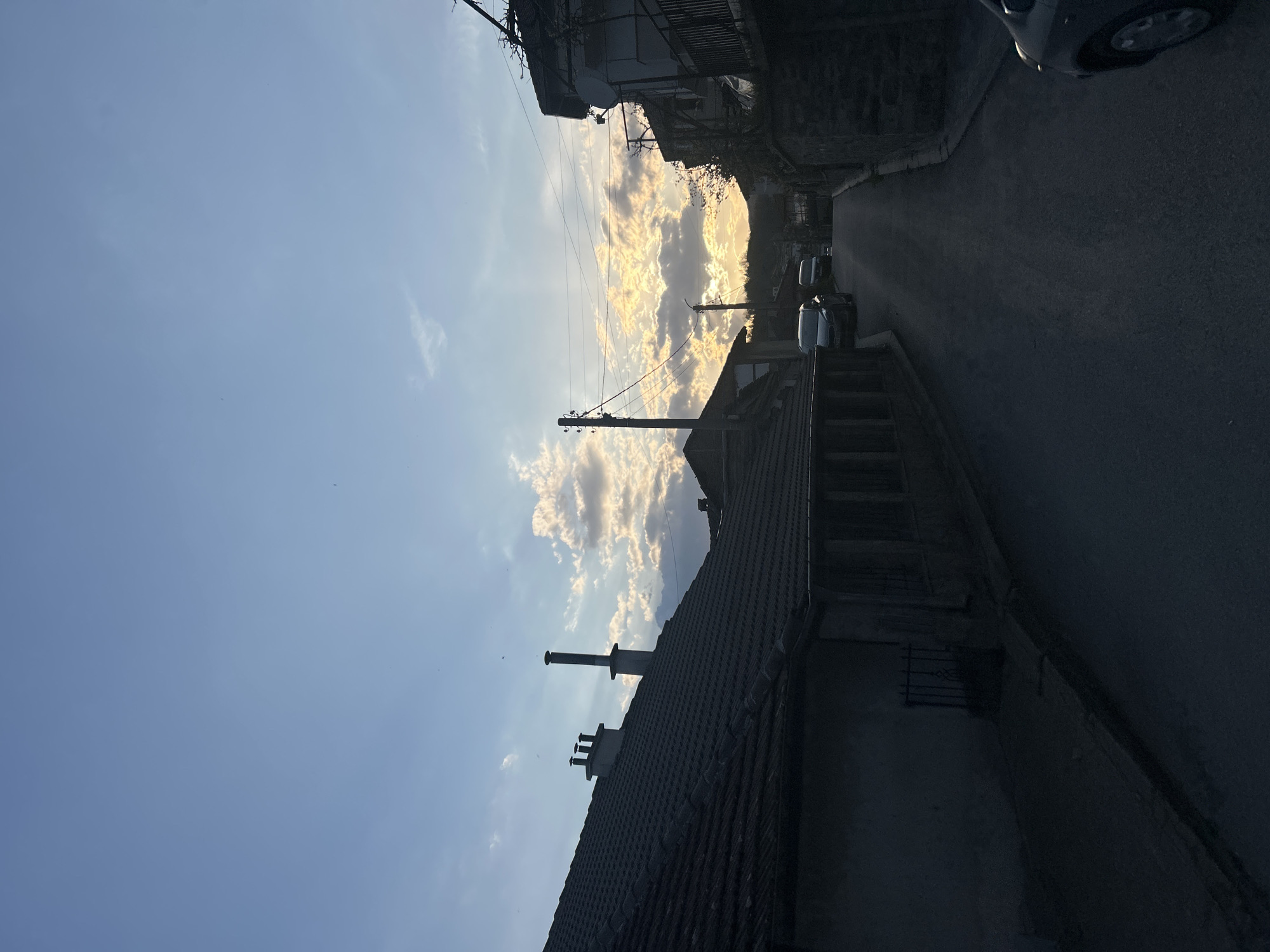 The image captures a picturesque street scene in Zlatograd, Bulgaria, at either sunrise or sunset. The sky dominates the upper portion of the frame, displaying a transition from clear, light blue on the far left to a vibrant display of glowing orange, yellow, and white clouds where the low-lying sun breaks through on the left. Small, dark specks, possibly birds, are scattered across the sky.
Below the dramatic sky, residential buildings with dark, tiled roofs line a narrow, paved street. The architecture suggests a traditional European town. On the bottom side of the image, the rooftops of closer buildings are visible in silhouette, featuring two prominent chimneys on one structure and a metal grate on a wall. The street, appearing dark and asphalt-paved, stretches into the distance on the right, with several parked cars partially visible along its edge, also largely in shadow. A utility pole with overhead wires extends diagonally across the mid-ground, adding an element of urban infrastructure.
The scene is characterized by strong backlighting, which casts the buildings, street, and vehicles into deep silhouette, enhancing the warm, golden glow of the sunlit clouds. This contrast between light and shadow, combined with the tranquil setting, evokes a serene and contemplative mood. No people or visible text are present in the image, and no specific activity is taking place.