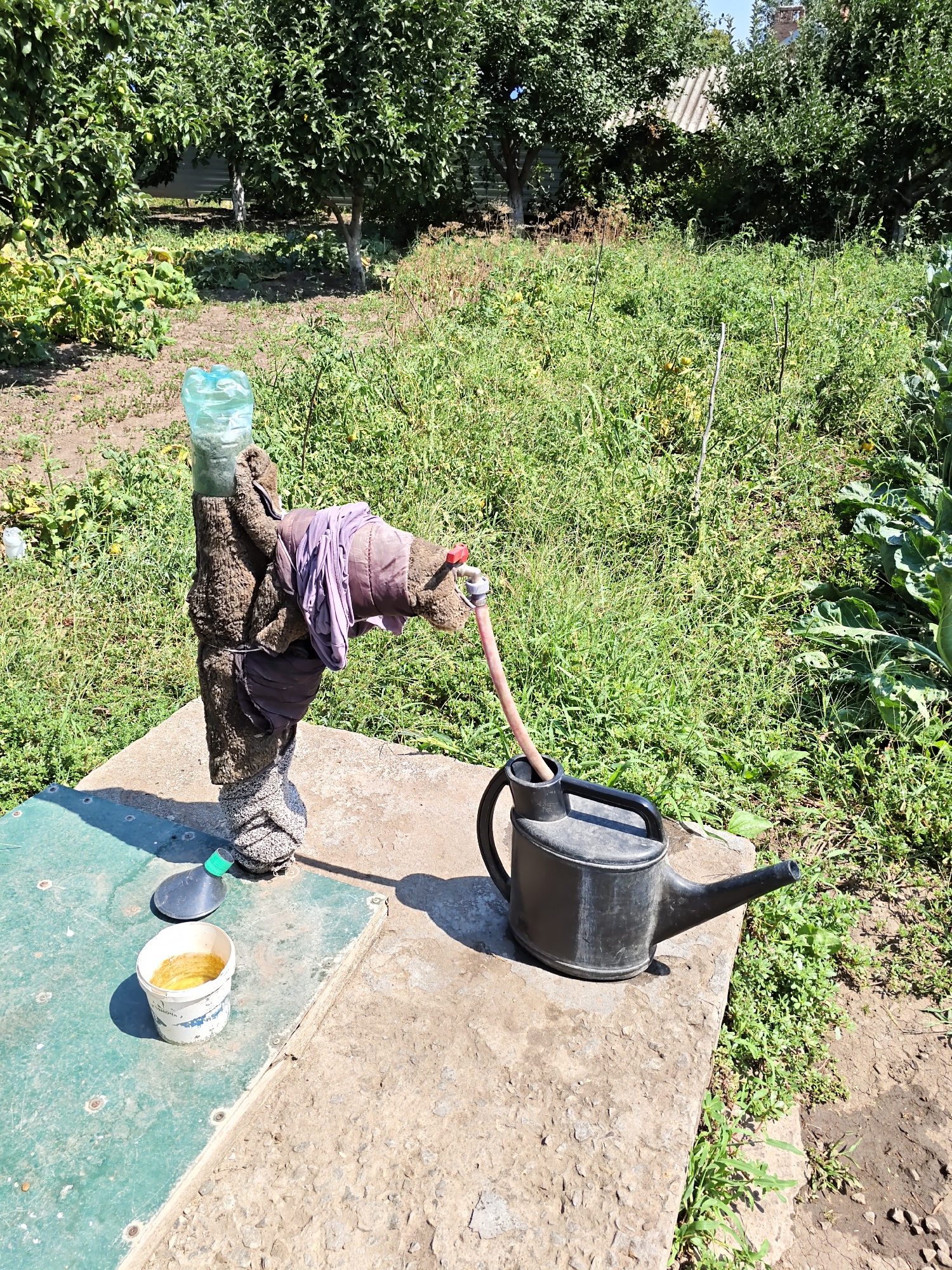 In the city of Nizhnedonskoy, Russia, an outdoor scene depicts a makeshift water faucet. The faucet is a vertical pipe wrapped in brown material, resembling a blanket, and secured with dark fabric strips. A clear plastic bottle is attached to the top of the pipe. A pink hose extends from a red faucet head on the side of the pipe, arching over and emptying into a black watering can positioned on a concrete platform. To the left of the faucet, a small white bucket with yellow liquid sits on a green surface. The background is filled with lush green vegetation, including trees and leafy plants, suggesting a garden or rural setting under a clear blue sky.