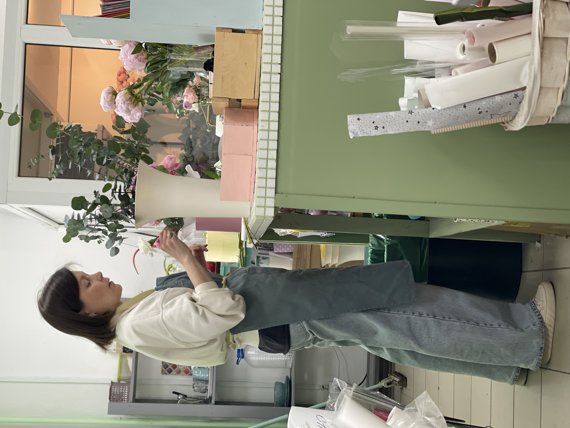 A medium shot captures a woman standing in a flower shop in Unknown, Unknown. She is fair-skinned with dark hair, wearing a white sweatshirt and light blue jeans with an apron. She is holding a flower, possibly arranging an arrangement.
Behind her, a light-green shelving unit holds transparent and white paper rolls, some with star patterns. Next to her is a counter where flowers in a vase can be seen. The overall environment suggests a florist workspace.
