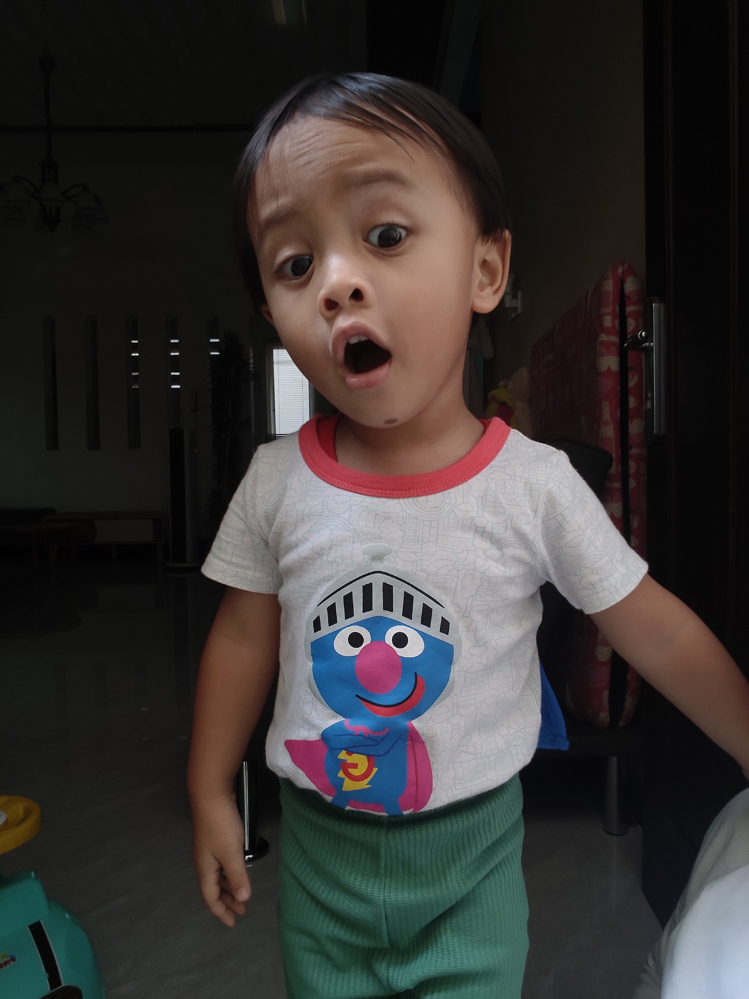 The image shows a young child, appearing to be between 3 and 5 years old, with dark hair and wide, surprised eyes. The child is wearing a white t-shirt with a graphic print of a blue character with a pink nose and a yellow and blue cape. The child is also wearing green shorts. The child's mouth is open in an expression of surprise or excitement. The background is dimly lit and appears to be an indoor setting, possibly a doorway or entrance area. Some furniture and architectural details are visible in the background, suggesting a residential or commercial space. The lighting suggests the photo was taken indoors, possibly near a window or open door, with the primary light source coming from the front. There are no specific visual cues in the image that directly indicate the location is Pemalang, Indonesia. The description is solely based on the visual content of the photograph.