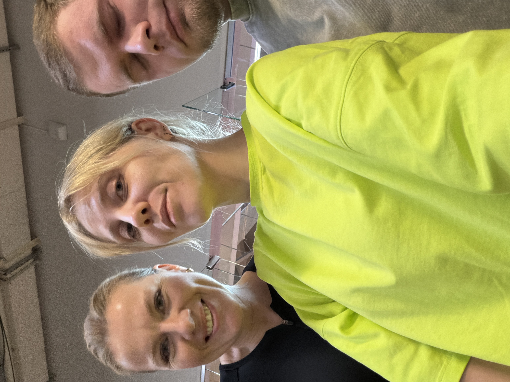 The image depicts three individuals in a low-angle selfie. At the bottom is a woman with light blonde hair pulled back, wearing a black top and smiling. Above her is another woman, also with light blonde hair, wearing a bright lime green shirt and a neutral expression. At the top of the frame is a man with short brown hair and a beard, his eyes closed. The background appears to be a room with a light-colored ceiling and walls. The location is identified as Chaikovsky, Russia.