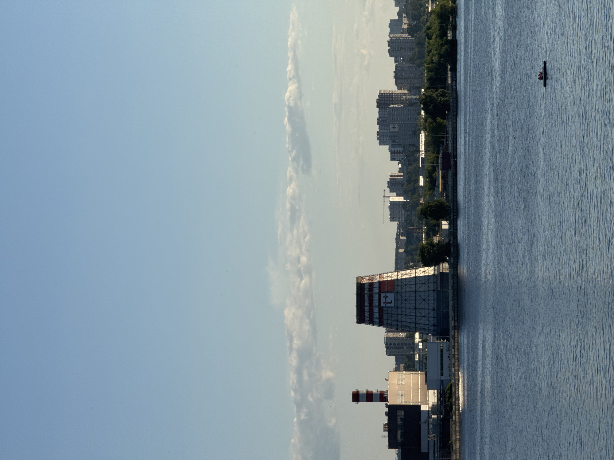 The image presents a wide-angle view across a body of water, identified as the Izhevsk Pond (Izhevsky Prud), towards the city of Izhevsk, Russia. The upper two-thirds of the frame consists of a clear, light blue sky with a horizontal band of white cumulus clouds at the mid-horizon.
The lower third features the dark blue water of the pond, exhibiting minor ripples. In the mid-ground, on the far bank, an industrial complex is prominent. This complex includes a tall, red and white striped chimney and a large, hyperboloid cooling tower. The cooling tower's upper section is red with a white lattice structure, displaying a white "Т+" logo and the Cyrillic text "ИЖЕВСКАЯ ТЭЦ-1," indicating the Izhevsk Thermal Power Plant No. 1. Adjacent to these structures are several industrial buildings, characterized by dark and light-colored facades.
Beyond the industrial structures, the background reveals the Izhevsk cityscape, composed of numerous multi-story buildings, appearing as residential or commercial blocks. A single construction crane is visible on the left side of the cityscape. Patches of green vegetation, including trees, are present along the shoreline and within the urban backdrop.
In the lower right portion of the water, a small, white pedal boat is observed, containing two individuals who appear to be propelling the boat. The scene is illuminated by natural daylight, suggesting late afternoon or early evening.