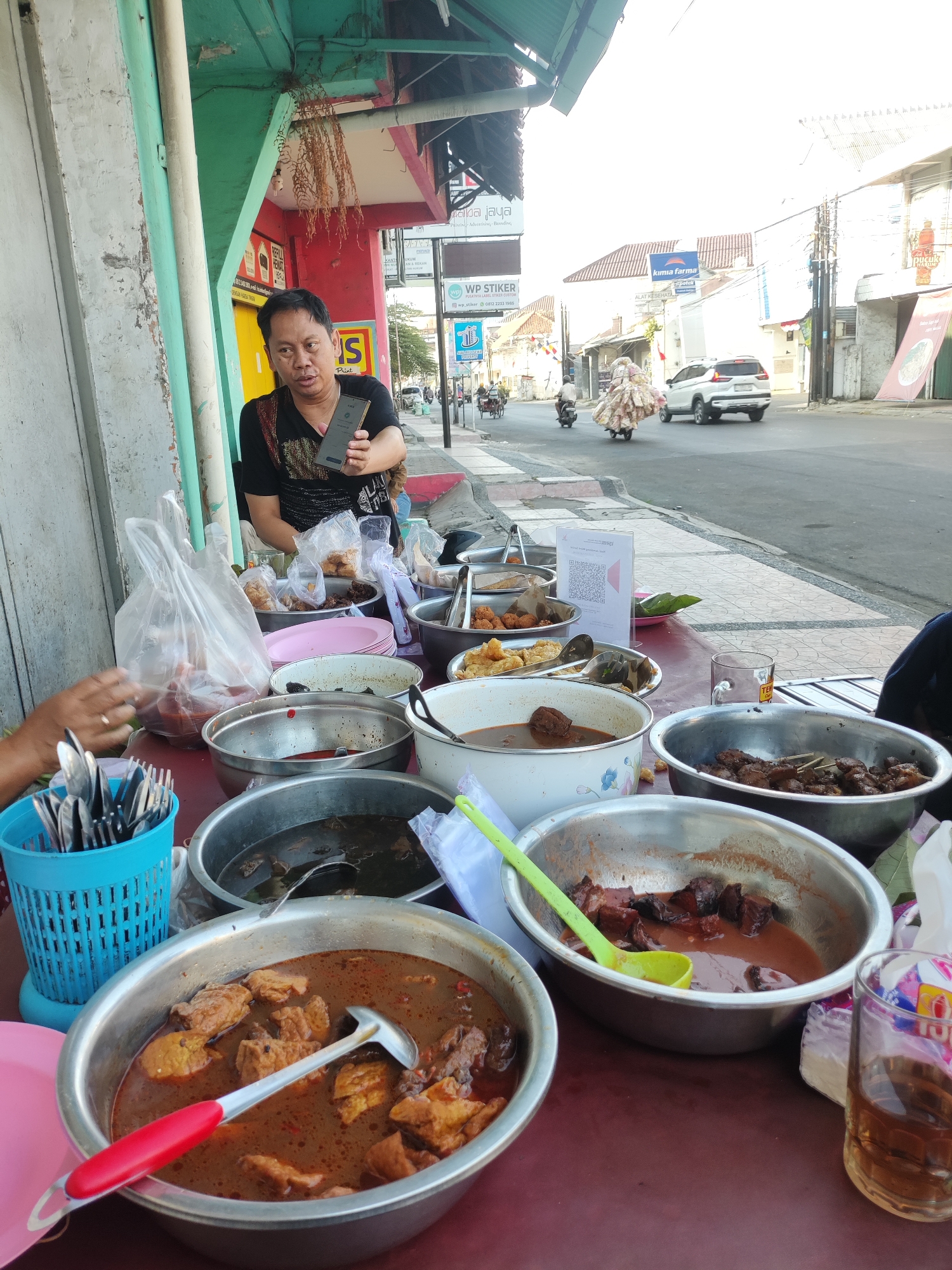 In Cirebon, Indonesia, a street food vendor is captured at a stall on a sidewalk, displaying an assortment of dishes in metal bowls. The vendor, a middle-aged man wearing a black t-shirt, is holding up a mobile phone, possibly for payment or communication. The food appears to be a variety of stews and fried items, with some packaged in plastic bags. In the background, a street scene unfolds with cars, motorcycles, and pedestrians, indicating an urban setting. The stall is positioned beside a building with a green and red facade, and a street sign for "WP STIKER" is visible. The overall scene depicts a typical roadside eatery in Indonesia, emphasizing the local culinary culture and daily commerce.