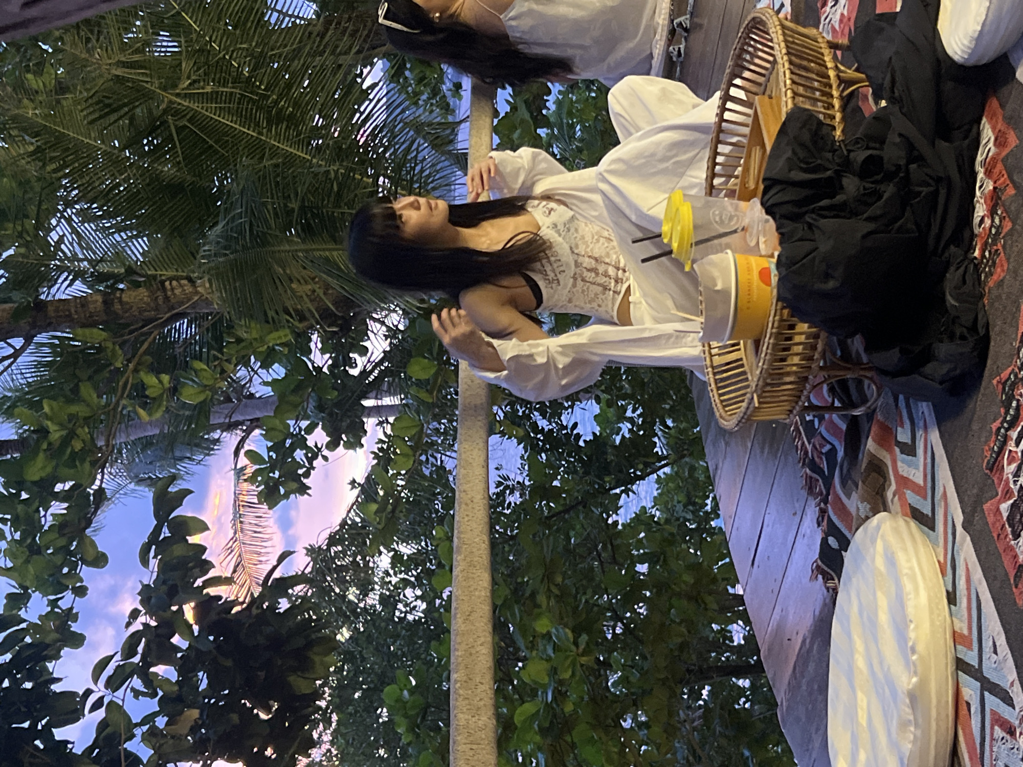 In Karon, Thailand, on a wooden deck overlooking the ocean and lush greenery, two women are seated. The woman in the foreground, with black hair and bangs, is wearing a white lace bralette and flowy white pants and top. She is seated cross-legged, looking to her right, with her hands near her chest. In front of her is a woven tray with a yellow cup, a clear cup with a straw, and a yellow container. To her left are two white cushions on a patterned rug. The second woman, in the background, is wearing a white top and sunglasses. She is also looking to her right. The sky is a twilight purple and pink.