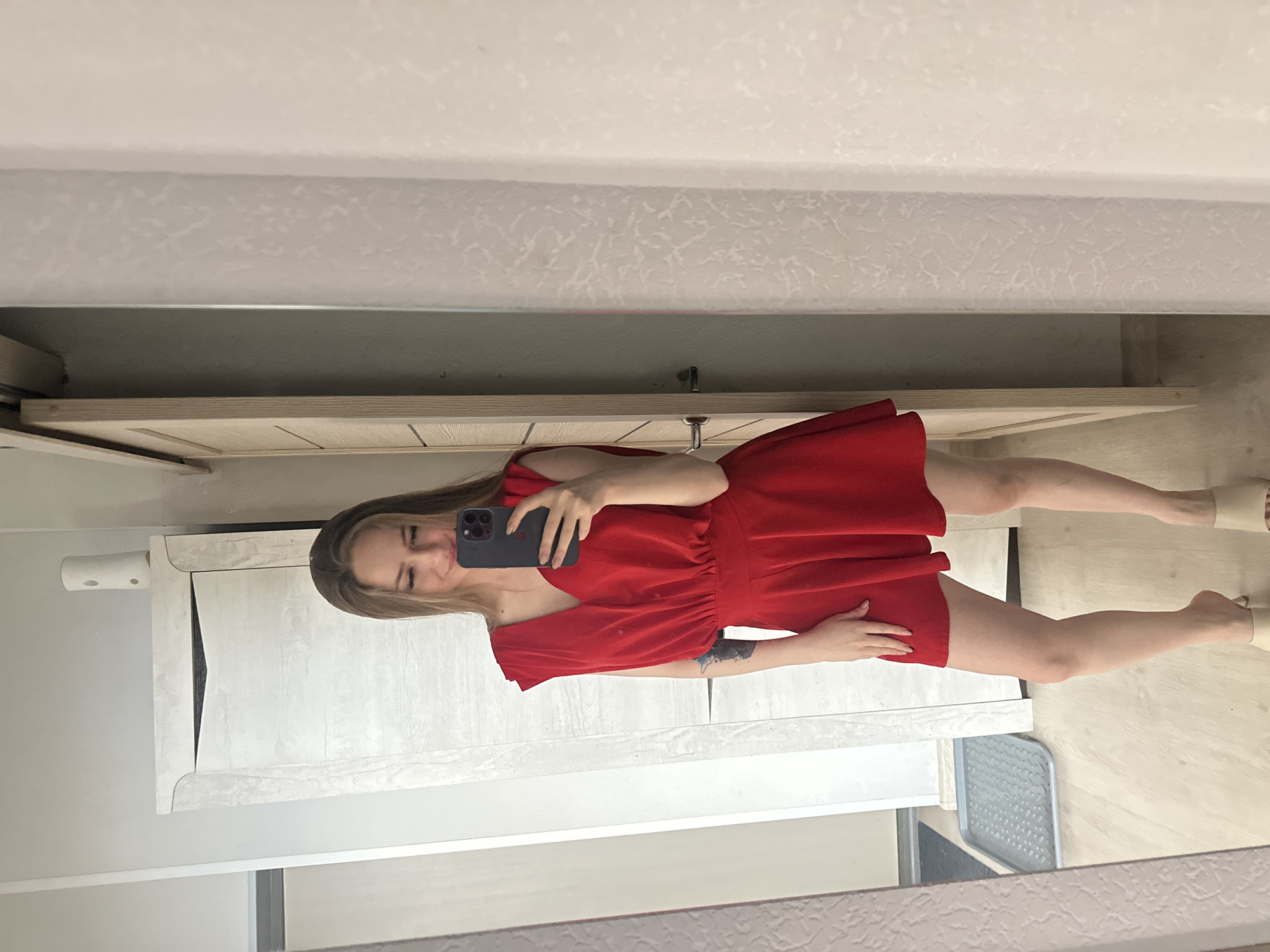 A young woman with long blonde hair, wearing a red v-neck dress and beige mules, stands in front of a mirror, taking a selfie with a black iPhone. She has a tattoo on her left arm. The background shows a wooden closet door, a white dresser with a cylindrical object on top, and a white textured wall. The floor appears to be light-colored wood. The setting is indoors, likely a bedroom or dressing area.