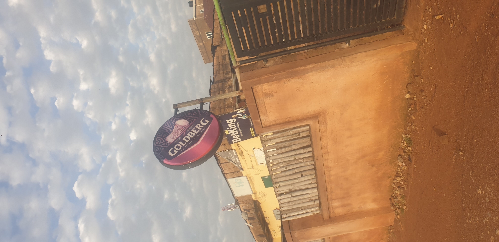 This image depicts an outdoor scene in Jos, Nigeria. A large, circular, pink and purple illuminated sign for "GOLDBERG PREMIUM LAGER BEER" is prominently displayed against a cloudy sky. Below the Goldberg sign, a smaller rectangular sign for "BetKing" is visible. This BetKing sign features an image of a man in a suit and the slogan "Bet Feeling. That's Betting." The background includes the rooftops of buildings, some with corrugated iron, and a fence made of bamboo poles. A metal gate with horizontal bars is also present on the right side of the image. The foreground shows a dirt road with scattered pebbles and rocks. The overall impression is of a commercial area or a street scene in a city.
