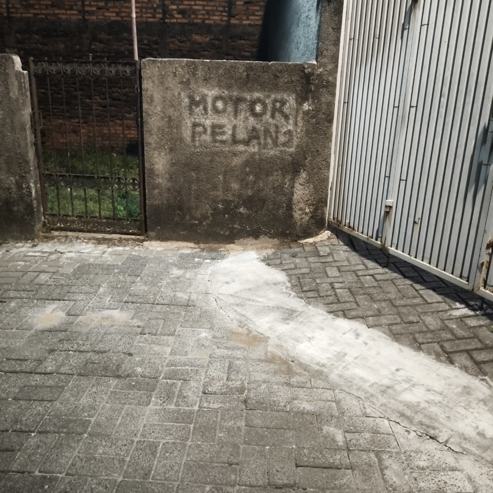 The image depicts an outdoor scene, possibly a street or alleyway, in South Jakarta, Indonesia. The foreground features a paved ground of gray interlocking bricks, with some areas showing discoloration and cracks, suggesting recent water spillage or wear. To the left, a metal gate with intricate, decorative patterns stands partially open, revealing lush green vegetation behind it. Adjacent to the gate is a rough, textured concrete wall. In the center of this wall, the words "MOTOR PELAN" are stenciled or painted in a distressed, rough font, with the letters appearing worn and somewhat faded. To the right of the wall, a tall, corrugated metal gate, painted in a light gray or silver hue, dominates the background. The overall lighting suggests it might be dusk or a dimly lit area. The environment appears to be residential or commercial, with a brick wall visible further in the background. There are no people or animals visible in the image.