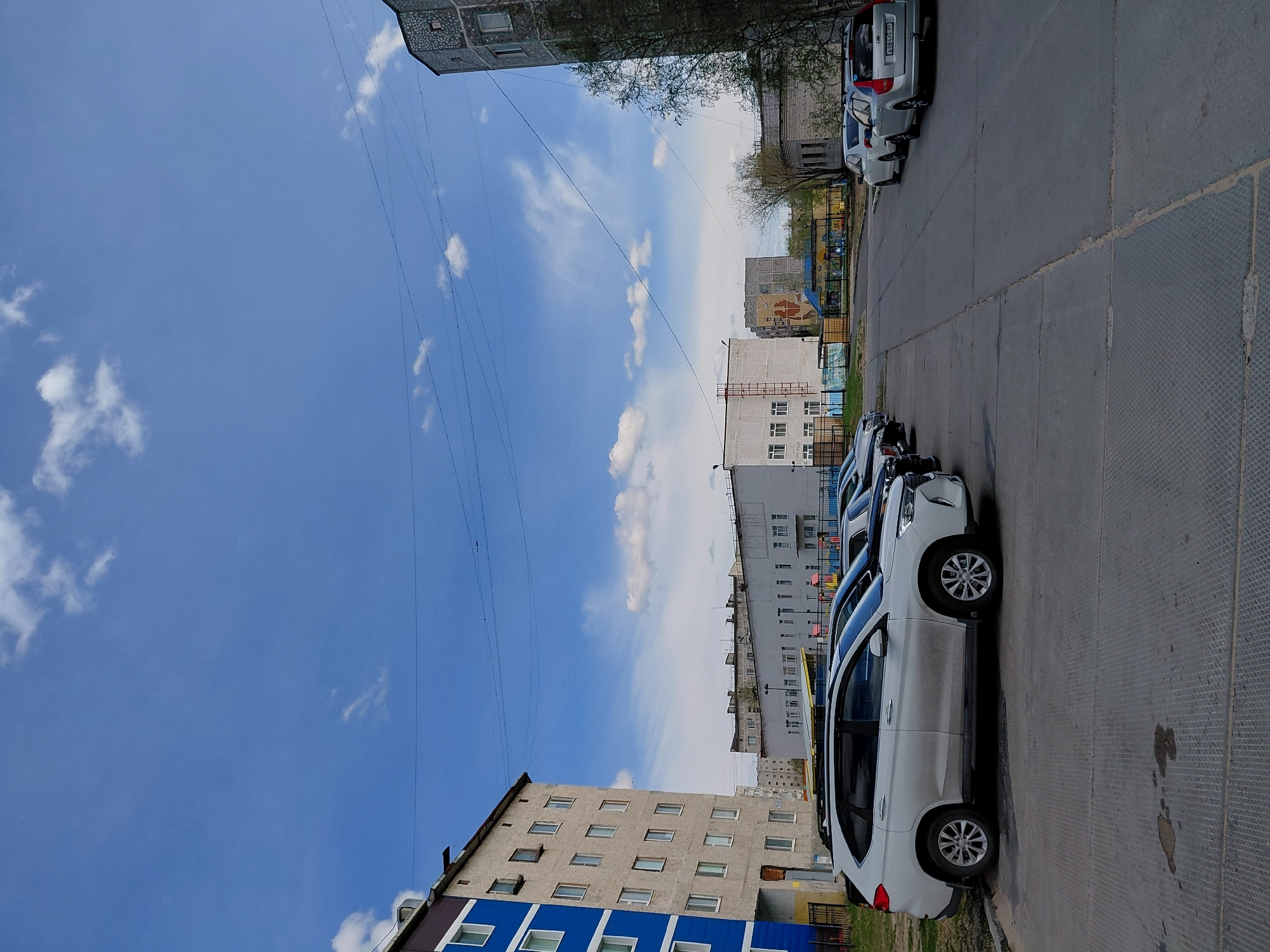 The image captures an outdoor urban scene in Nadym, Russia, showing a street or parking area bordered by multi-story residential buildings under a bright sky. The perspective is slightly elevated, looking down along a line of parked cars.
In the foreground, a white SUV is prominently parked on a paved surface, with its wheels visible. Several other cars are parked behind it, extending into the mid-ground. One silver car in the distance has a visible license plate: "T082EP 186," which corresponds to the Yamal-Nenets Autonomous Okrug, where Nadym is located. The pavement shows seams and some textured sections, with a few dark stains visible on the ground in the immediate foreground.
To the left, a multi-story apartment building with a distinctive blue lower facade and numerous windows rises. Further back, another older-looking building is partially visible. On the right side of the image, more buildings are present, with sparse trees suggesting a time of year when foliage is minimal, possibly early spring or late autumn. A colorful structure, likely part of a children's playground, is visible in the mid-ground between the buildings and cars.
The sky is a clear, vibrant blue with scattered white, wispy clouds, indicating good weather. Numerous thin power or communication lines crisscross high in the sky. The overall scene depicts a mundane, everyday moment in a residential area, under bright daylight conditions. No specific human activity is evident; the focus is on the urban landscape and parked vehicles.