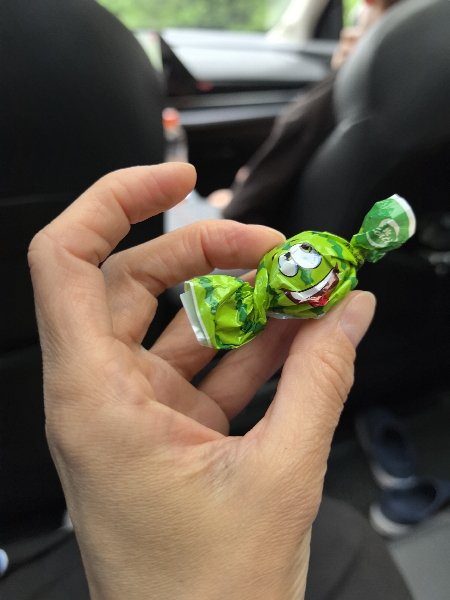 A person's hand holds a green candy wrapper with a cartoon face. The wrapper is crinkled and appears to be made of foil or plastic, with a drawing of a watermelon-like face with large eyes and a wide, smiling mouth showing teeth and a red tongue. The background is blurred, showing the interior of a vehicle with dark seats and some light in the distance. The scene suggests a casual moment, possibly during a car ride. The context of Oktyabr'skii, Russia, is not discernible from the visual elements of the image itself.