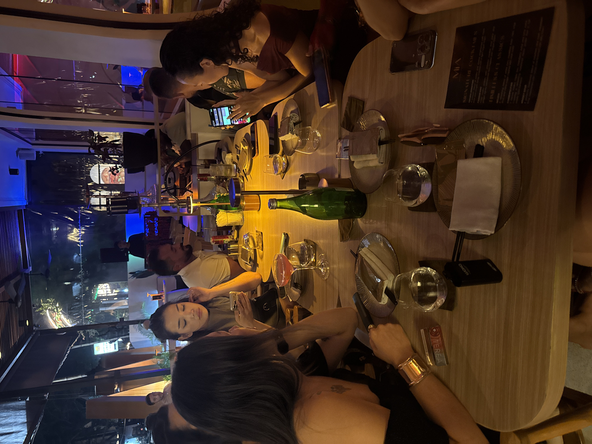 The image shows a group of people dining at Mia Restaurant in North Kuta, Indonesia. Diners are seated around a long table, some are looking at their phones. The table is set with plates, silverware, glasses, and drinks. A green bottle is also on the table. The setting is outdoors with views of the surrounding area and a cityscape in the background. The image is taken at night.