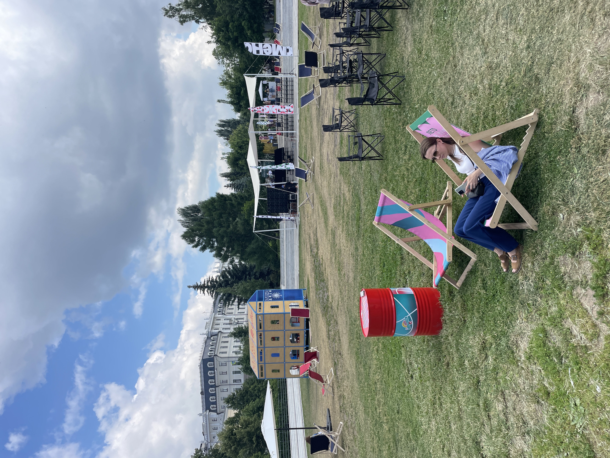 In Kazan, Russia, a woman relaxes on a striped deck chair in a park. She wears sunglasses, a white tank top, and blue pants, holding a smartphone. The park features a red barrel, other empty deck chairs, and folding chairs arranged on the grass. In the background, a stage with white tents and banners is visible, with trees and a large building in the distance. The sky is partly cloudy.