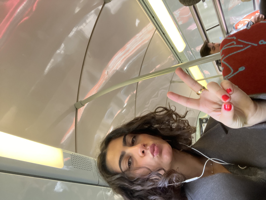 The image depicts the interior of a public transit vehicle, such as a train or metro car. The specific location in the city of Torcy, France, cannot be determined from the visual information provided.
In the foreground, a young woman with dark, curly, shoulder-length hair looks directly at the camera with her lips pursed in a pout. She is wearing a grey overcoat, a delicate necklace with a small pendant, and white wired earbuds. Her left hand is raised to her left, forming a peace sign (V-sign) with her index and middle fingers extended. Her fingernails are painted red, and a plain gold band ring is visible on her left ring finger.
Partially visible in the background, to the right of the woman, is a young man with dark hair, seated and looking downward or away from the camera. The vehicle's interior features a white, curved ceiling with embedded fluorescent strip lighting and a rectangular ventilation grille. Light-colored, curved support poles are present. Seating visible in the background is upholstered in red fabric with light blue, abstract blob-like patterns.