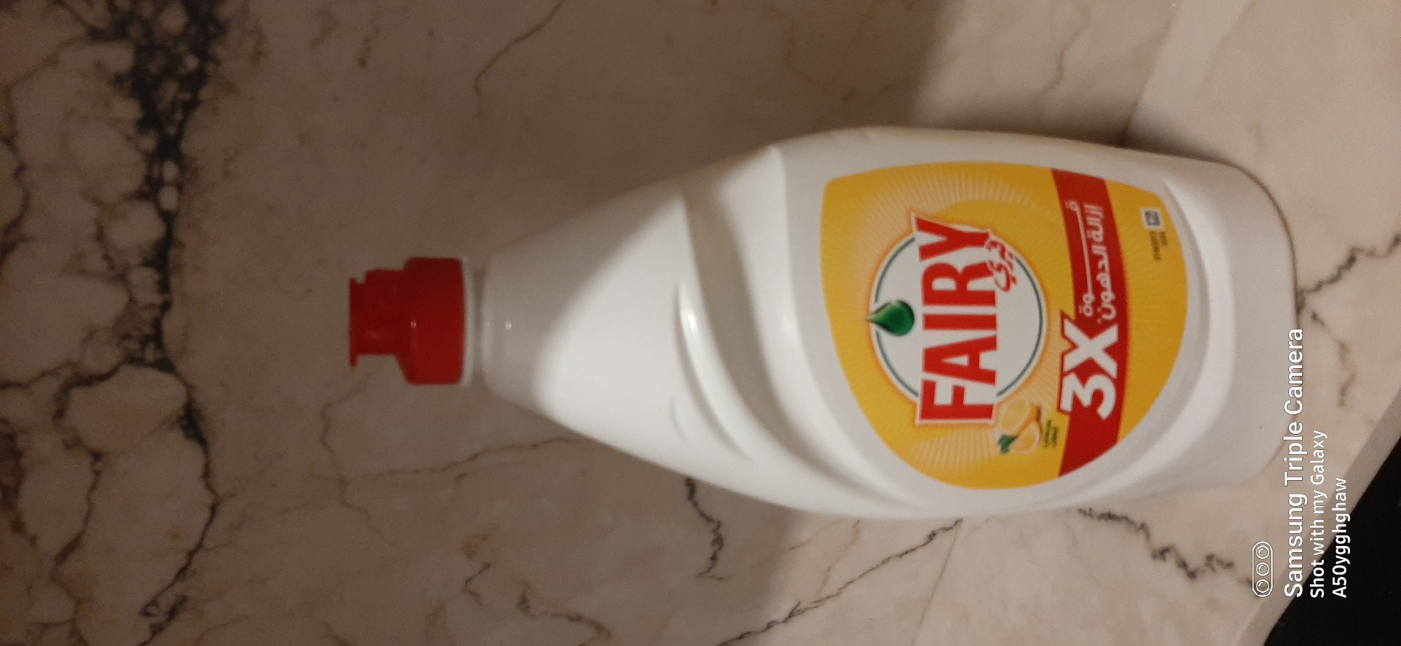 A white bottle of Fairy dish soap with a red cap stands on a marble countertop. The bottle has a yellow label with red lettering that says "FAIRY" and "3X". The background is a marble wall with brown and white veining. There is a "Samsung Triple Camera" watermark at the bottom left of the image. The scene appears to be an indoor kitchen setting.