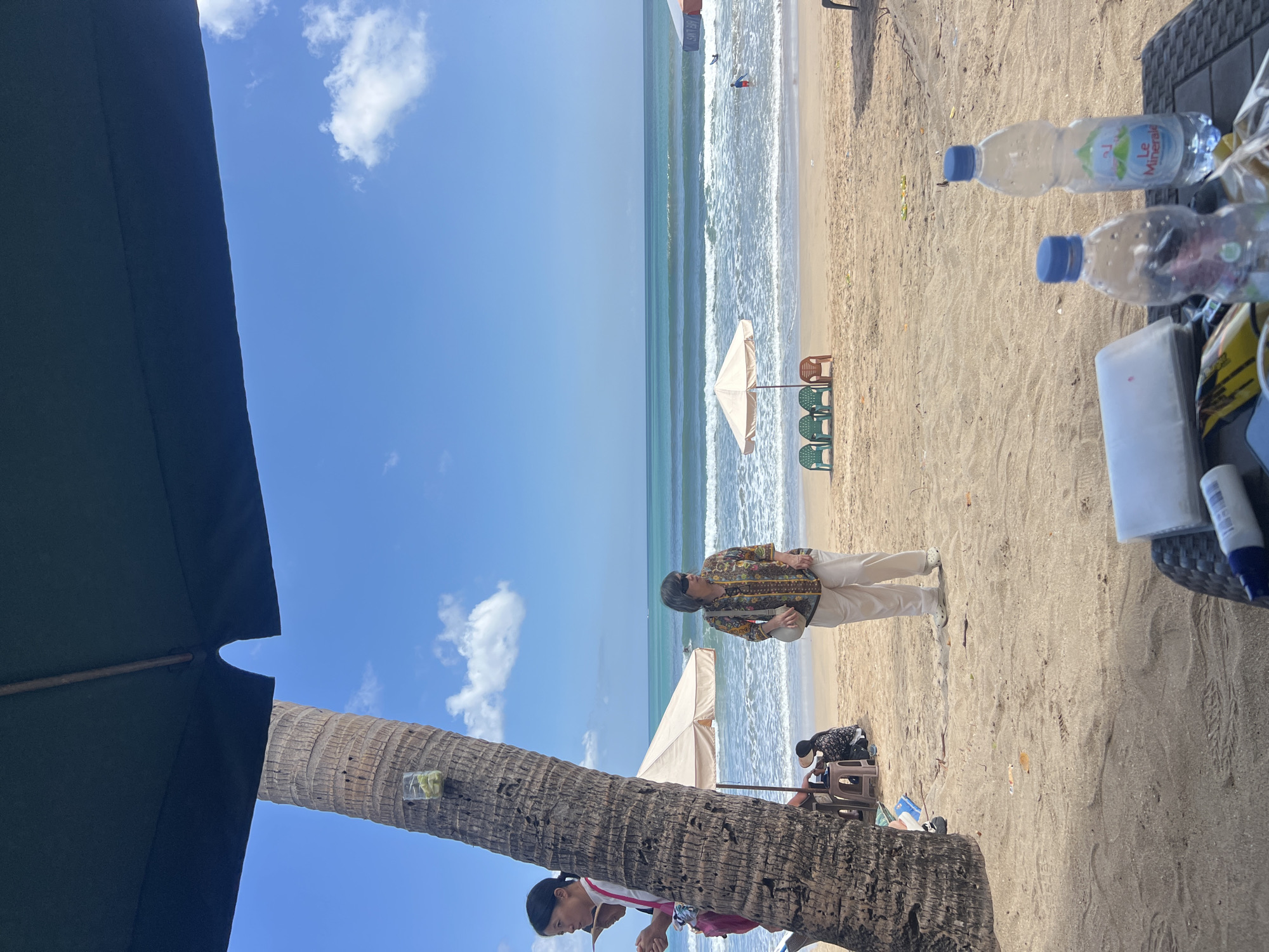 This image depicts a beach scene in Kuta, Indonesia, under a bright blue sky with scattered clouds. The foreground is dominated by a large palm tree on the left and a table with two plastic water bottles and other items in the lower right corner. A dark umbrella partially obscures the top of the image.
In the mid-ground, a woman wearing sunglasses and a patterned shirt stands on the sandy beach, facing towards the ocean. Behind her, a beige beach umbrella stands over a few plastic chairs. Further out, the turquoise ocean features gentle waves, with a few people visible in the water.
The sand is a light brown color, with footprints and scattered debris. The overall atmosphere suggests a leisurely day at the beach.