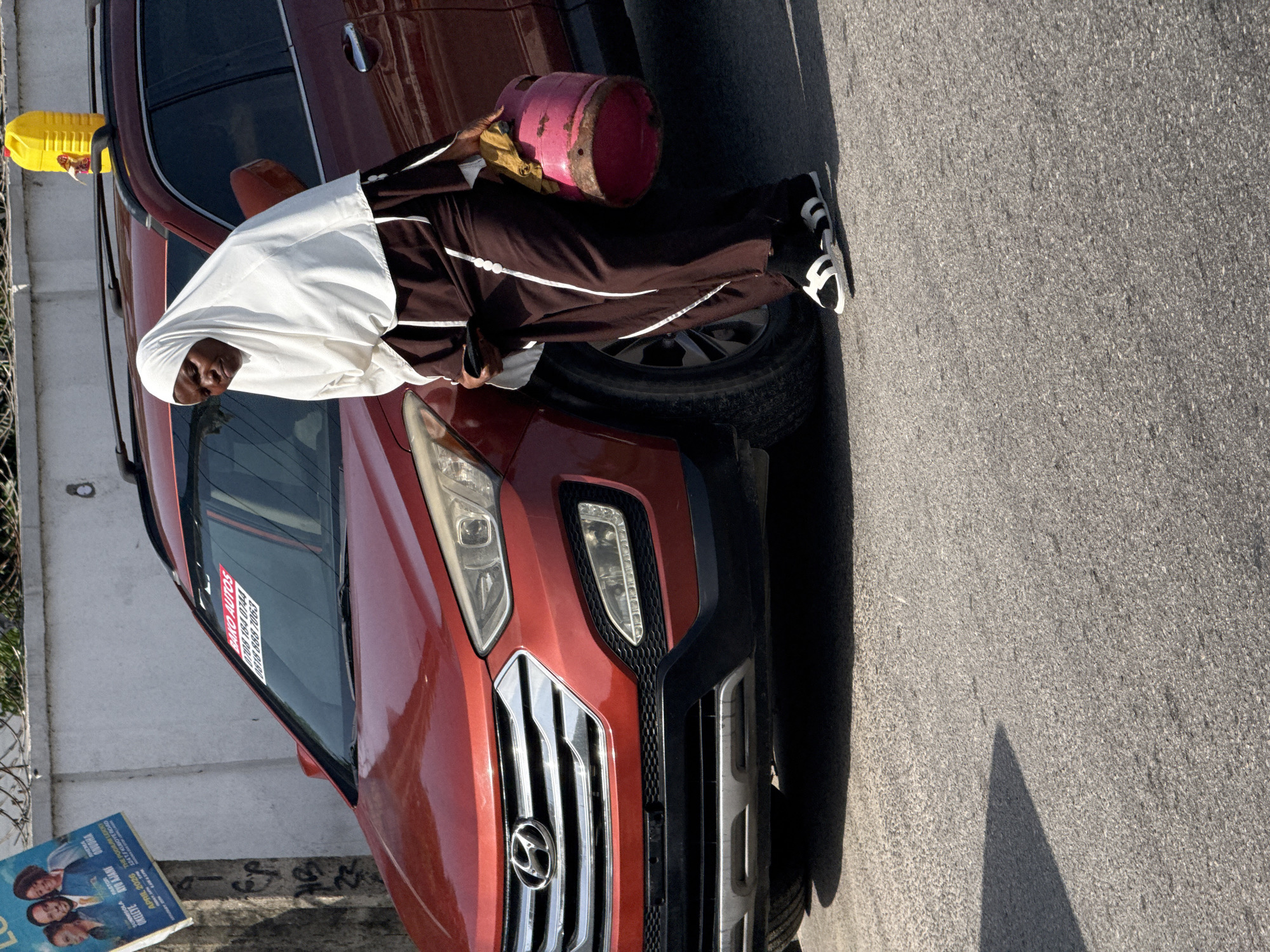 An elderly person, presumably female and wearing a white head covering and brown tracksuit, is near a red SUV in an urban setting. She is holding a pink propane tank. The vehicle has a "BAKO AUTOS" sticker on the windshield with Nigerian phone numbers. A discarded newspaper is visible near the front of the vehicle with "SUNMISOLA" printed on it. The background suggests a street in Ado, Nigeria.