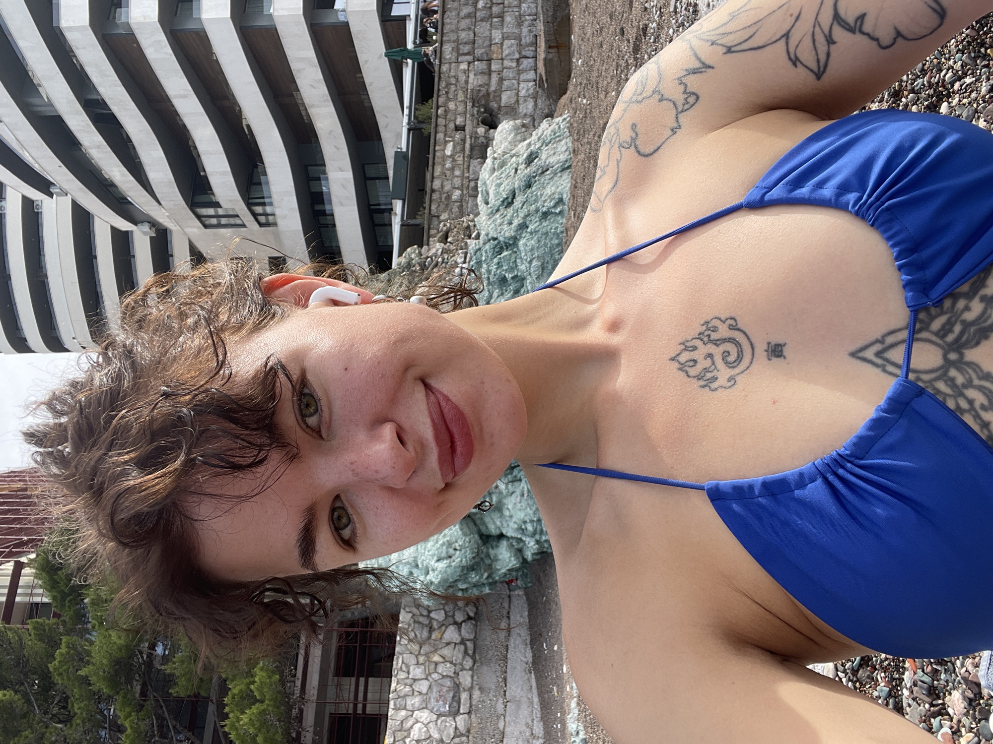 A close-up selfie captures a young woman outdoors, with the image rotated 90 degrees counter-clockwise. She has curly brown hair, striking green eyes, and a soft, closed-mouth smile, looking directly at the camera. She wears a royal blue string bikini top and white wireless earbuds are visible in her left ear.
Several tattoos adorn her body: a design resembling flames or waves with a small symbol is located on her sternum, while floral or leafy patterns are visible on her shoulders and arms.
The background shows a coastal scene. Immediately behind her head and shoulder, there's a light blue or turquoise rocky formation, an old stone wall, and green foliage. Further back, a modern building with distinctive black and white horizontal striped architecture stands prominently. The ground in the foreground appears to be a rocky shore or pebble beach. The bright, clear sunlight indicates it is daytime. The setting, with its mix of modern architecture, rocky coastline, and natural features, is consistent with coastal Rafailovići, Montenegro.