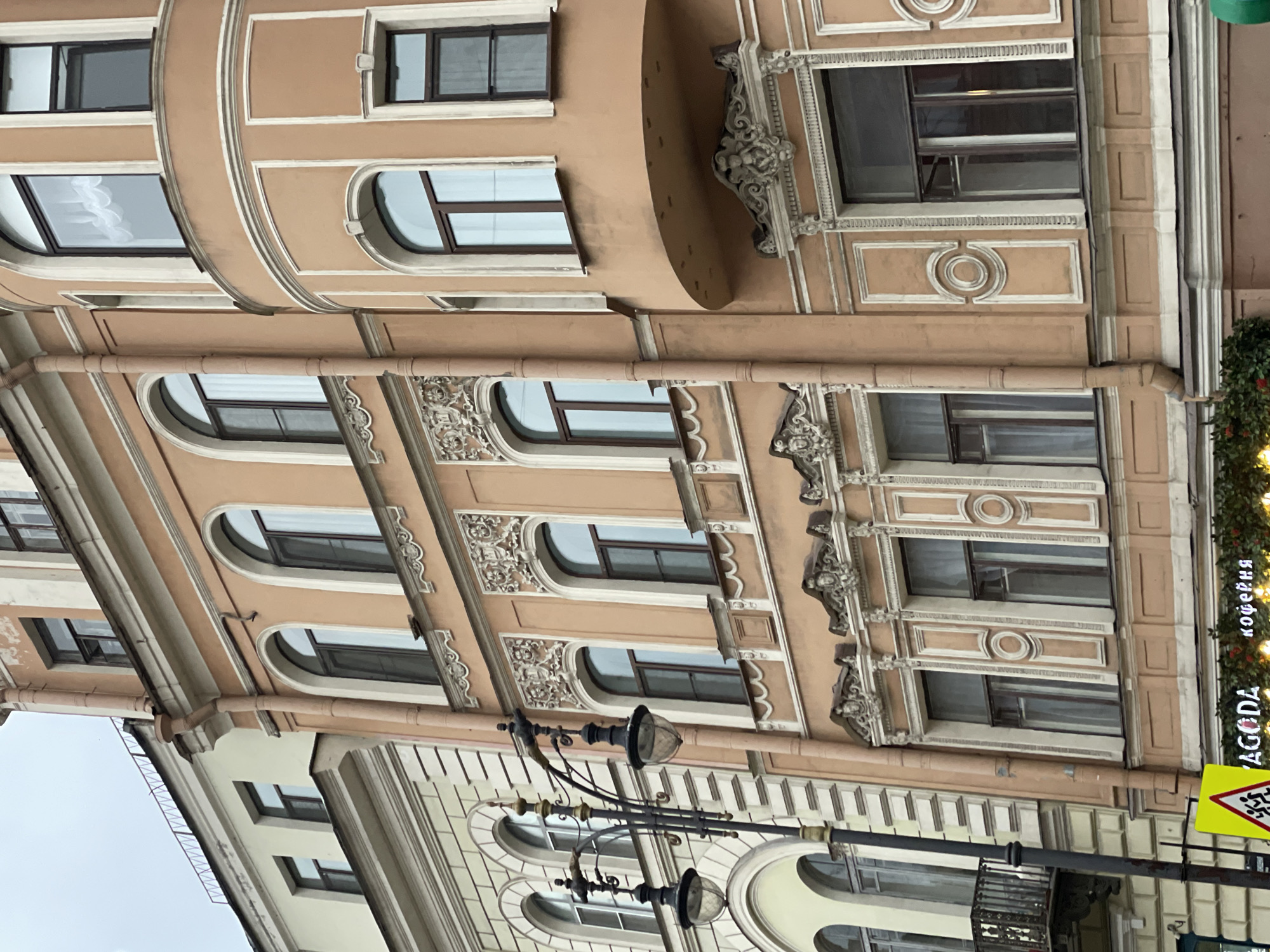The image shows the facade of a building, possibly an old apartment complex, in an urban setting, city and country unknown. The building is characterized by its ornate architectural details, including decorative moldings, arched windows, and a combination of rectangular and rounded window frames. The building has multiple stories, with windows spaced at regular intervals. The exterior is painted in a variety of colors, mainly beige and off-white. A vintage-style street lamp is situated near the bottom of the frame. A sign saying "AGODA" is visible on the right, as is a crosswalk sign. The photo is taken from an upward angle, focusing on the architectural features of the building. The sky appears overcast, and there are no people visible in the frame.