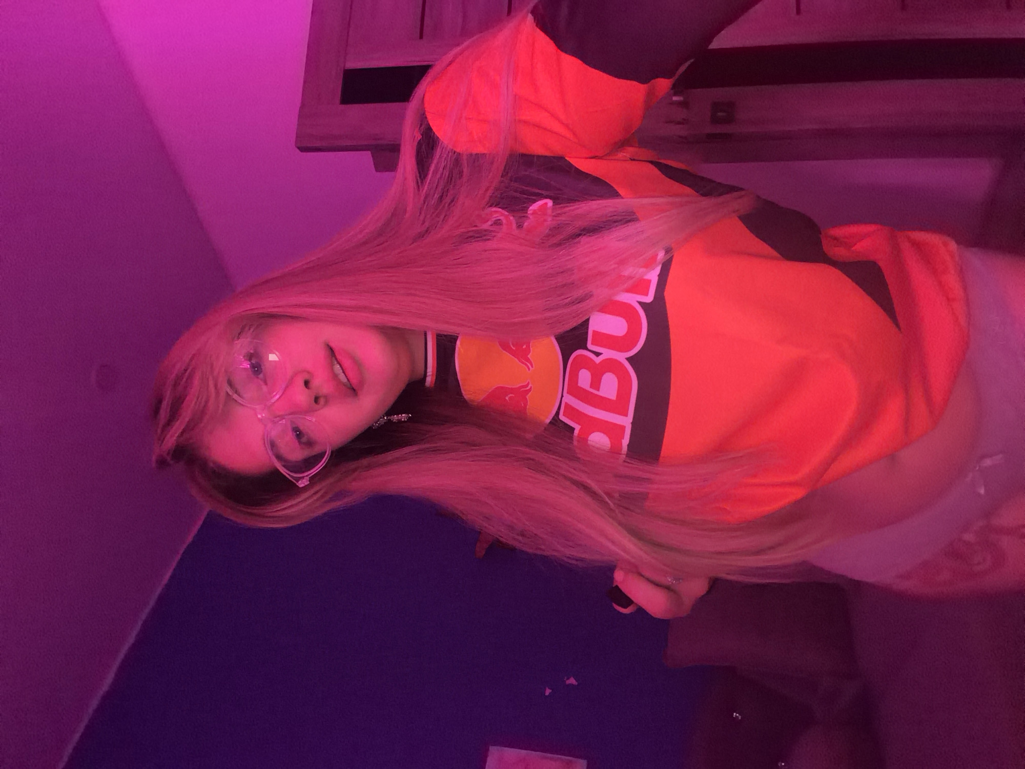 A young woman with long blonde hair is wearing clear-framed glasses and an orange, black, and yellow Red Bull shirt. She is indoors, possibly in a bedroom, under pink and purple lighting. A corner of a wooden table is visible above her. The wall is blue. The location is in the city of Bolshaya Shilna, Russia.