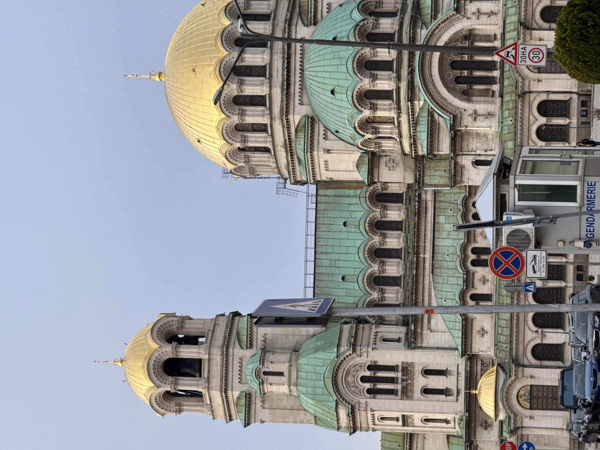 The image shows the Alexander Nevsky Cathedral in Sofia, Bulgaria. The architecture is characterized by golden domes and green roofs, with intricate stonework. Street signs and vehicles are visible in the foreground. A security booth labeled "Gendarmerie" is also present.