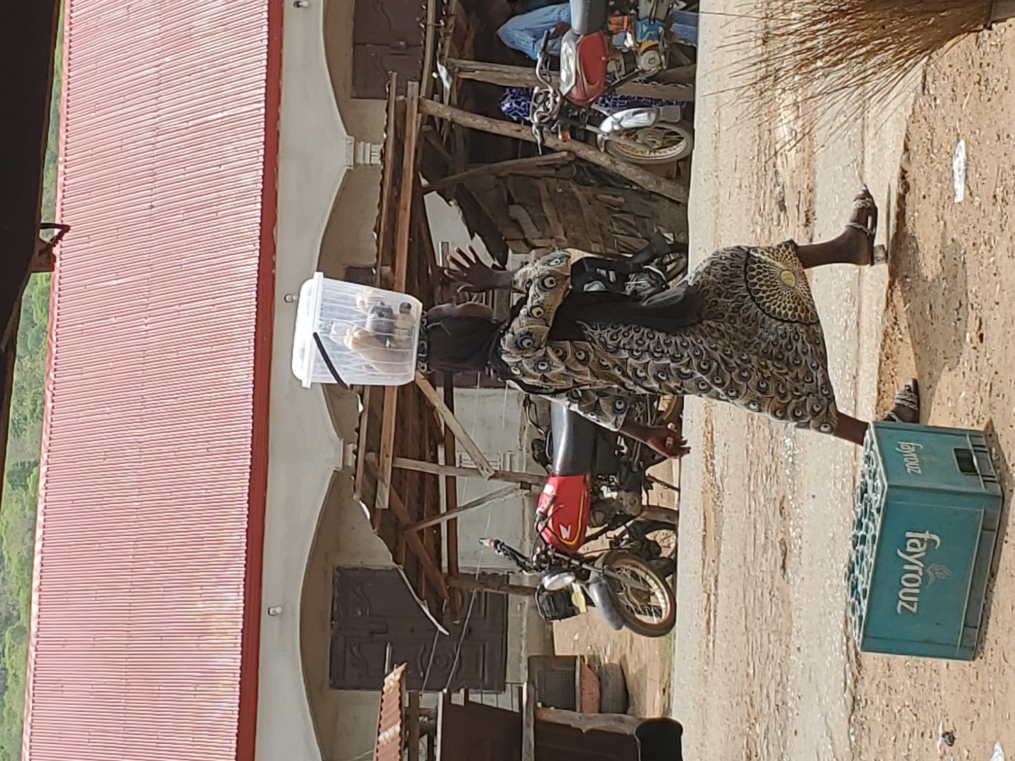 A woman in a patterned dress and a headscarf balances a clear plastic container on her head as she walks on a dirt road in Mararaba, Nigeria. She is passing by a building with a red corrugated metal roof and arched entryways. Behind her, two motorcycles are parked near wooden structures. A teal-colored Fayrouz soda crate sits on the ground beside her. The ground is uneven and dusty.