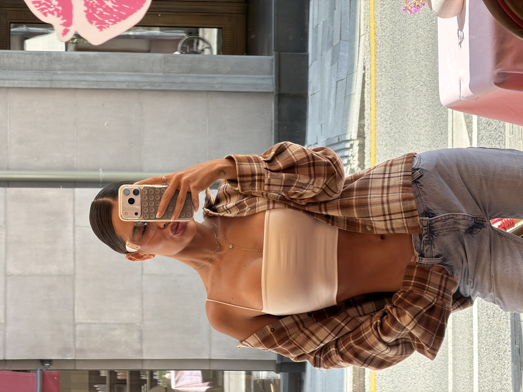A young woman is depicted taking a mirror selfie, facing the camera with a casual pose. She holds a smartphone, likely an iPhone given the camera configuration, which has a textured, grid-patterned case. Her dark hair is pulled back neatly, and she wears clear-framed glasses.
Her attire consists of a white, possibly strapless or spaghetti-strap, crop top that reveals her midriff, paired with an open, oversized plaid shirt in shades of brown, beige, and white. She also wears light-wash blue jeans. Accessories include several delicate necklaces, small earrings, and a ring on her right hand. A small, crescent moon tattoo is visible on her left forearm.
The setting appears to be indoors, likely in a shop or cafe with large reflective surfaces, in Moscow, Russia. The background is a reflection showing an urban street scene outside. This includes grey, concrete-like building facades and a yellow line on a paved surface, suggesting a street or sidewalk. Bright daylight illuminates the scene, implying it is mid-day or afternoon. A prominent pink decorative cutout, resembling a stylized heart or butterfly, is reflected in the upper part of the image, along with a pink rectangular object possibly a package or part of furniture, on the right. The image captures a contemporary, casual style. No visible text can be clearly identified.