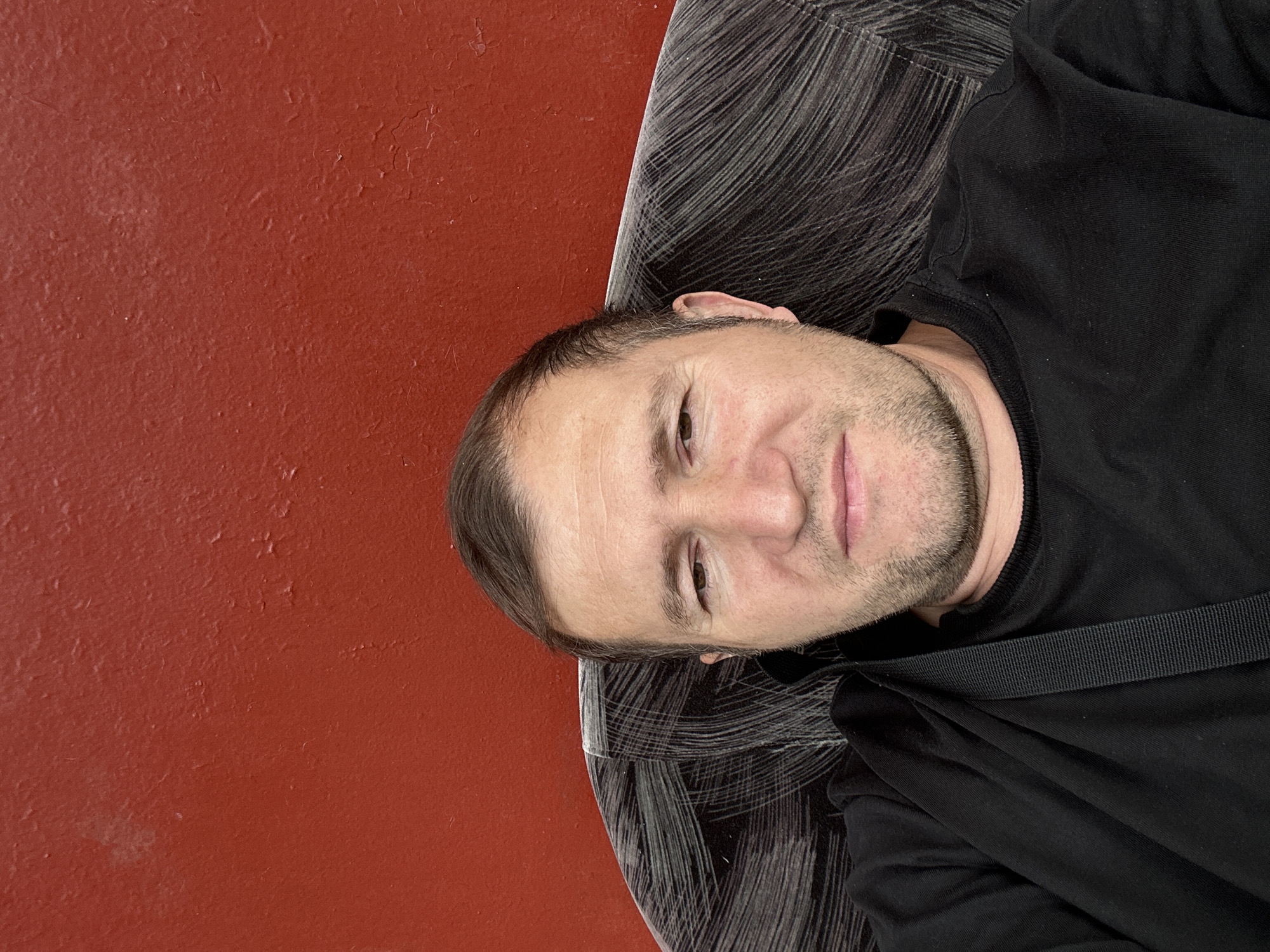 The media file is a close-up, top-down, selfie-style photograph of a light-skinned man with short brown hair and stubble. He is wearing a black t-shirt and appears to be lying down on a surface with a dark, patterned texture that resembles wood grain or brushed metal. The background is a solid, reddish-orange wall with some texture, suggesting it might be painted plaster. The lighting appears to be indoor, and the overall mood is casual and relaxed. The man's face is turned slightly towards the camera, and he is looking directly at the viewer with a neutral expression. His left eye is partially visible, and his lips are slightly parted. A black strap, possibly from a bag, is visible across his chest. The image is oriented horizontally.