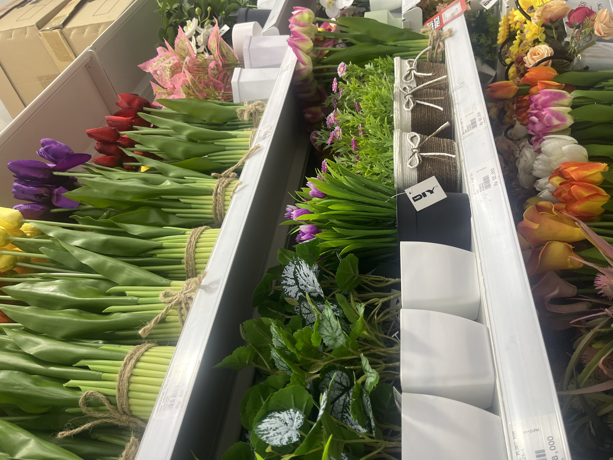 This image captures a vibrant display of artificial flowers and plants arranged on shelves within what appears to be a retail store, likely an aisle in a home decor or DIY shop. The scene is brightly lit, suggesting an indoor setting during business hours.
The primary subjects are numerous bundles of artificial tulips in a wide array of colors, including yellow, purple, red, pink, orange, and white. Many of these bundles are neatly tied with rustic twine, indicating they are ready for purchase. Alongside the tulips are various artificial green plants, some with simple green leaves, others featuring variegated green and white patterns. Smaller artificial flowering plants with purple blooms are also visible, some potted in white, minimalist rectangular containers.
Notable details include the orderly arrangement of products on white shelving units, creating a sense of abundance and variety. Several price tags are visible, displaying "Rp" (Indonesian Rupiah) amounts, such as "Rp 55,000", "Rp 68,500", and "Rp 88,000," confirming the location context of West Jakarta, Indonesia. One black tag clearly shows the "MR. DIY" brand logo. Other text includes product descriptions like "ARTIFICIAL PLANT" and product codes like "225 8046842" and "W02-06". Cardboard boxes are stacked on a higher shelf in the background. No people are visible, focusing attention entirely on the merchandise.